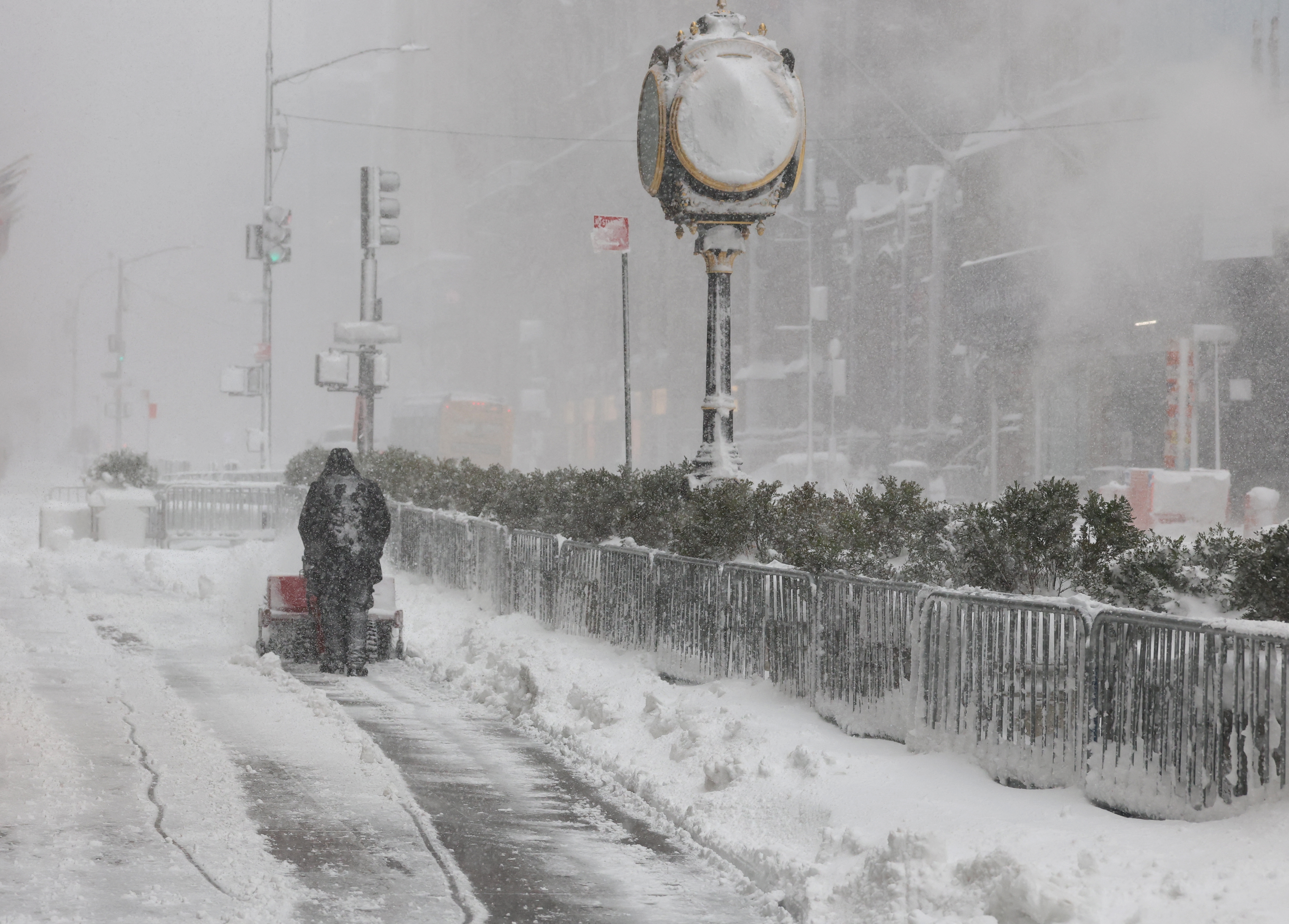 Person som går ned en snødekt bygate og dytter en snøfreser. Bygninger og en klokke er knapt synlige i kraftig snøfall