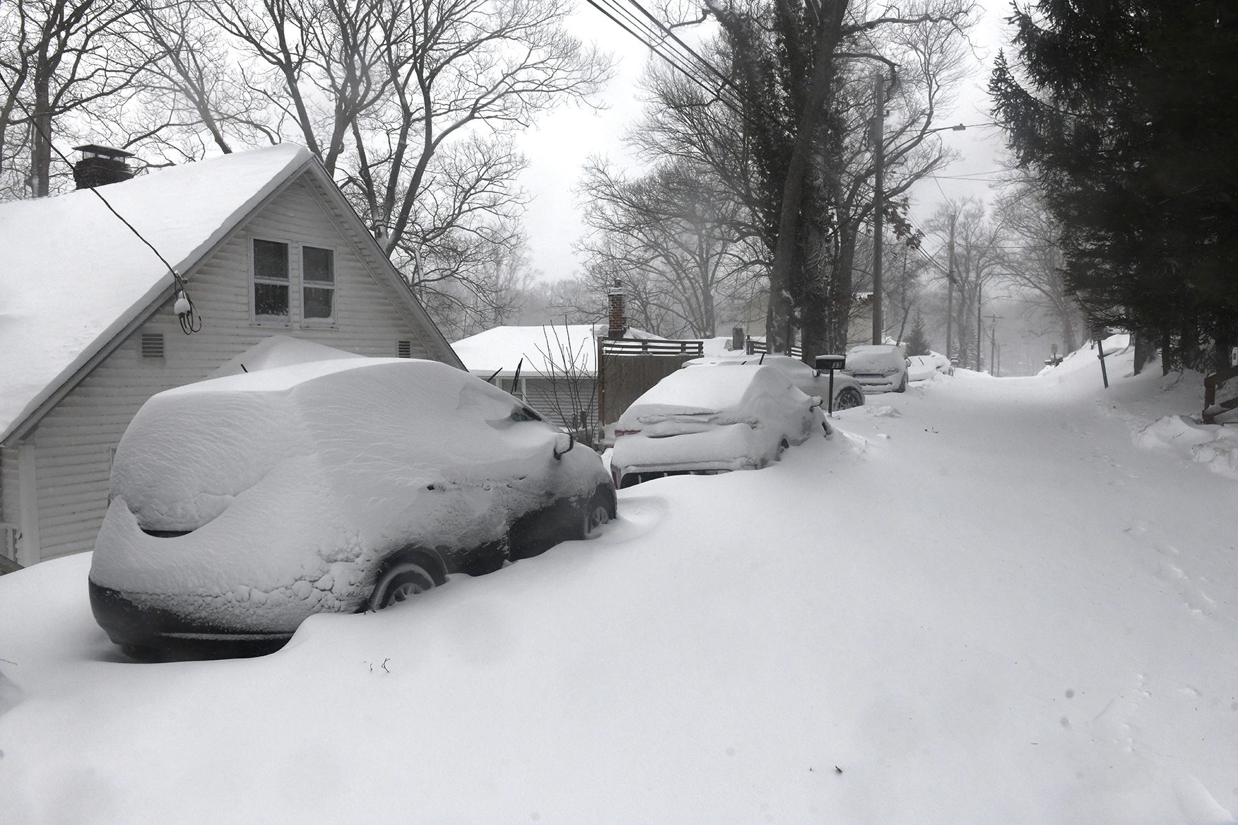 Snødekt gate med parkerte biler og et hus, trær i bakgrunnen, skaper en rolig vinterscene