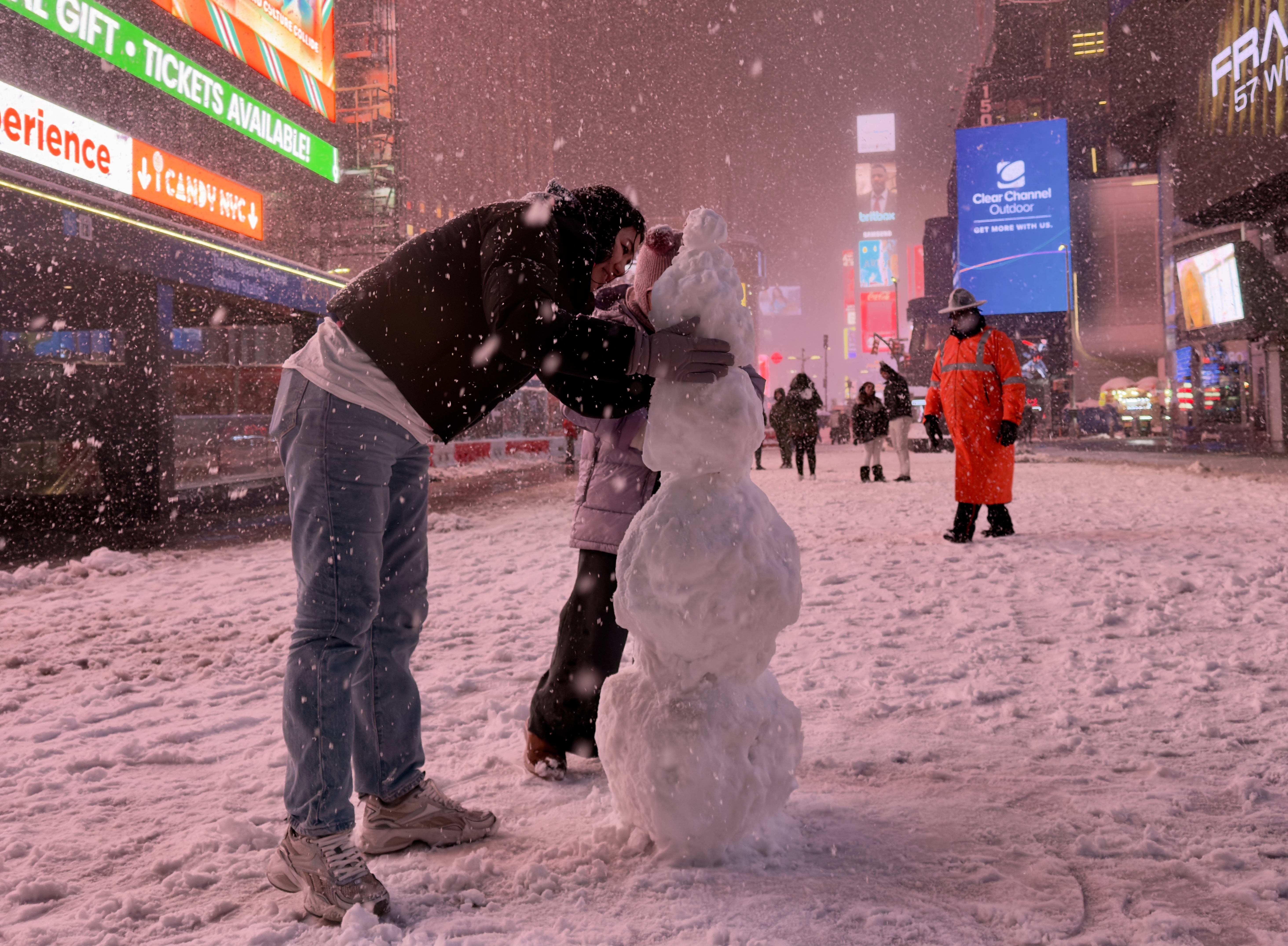 To personer bygger en snømann i en snødekt bygate mens snøen faller. Opplyste reklametavler og en person i oransje frakk er synlige i bakgrunnen