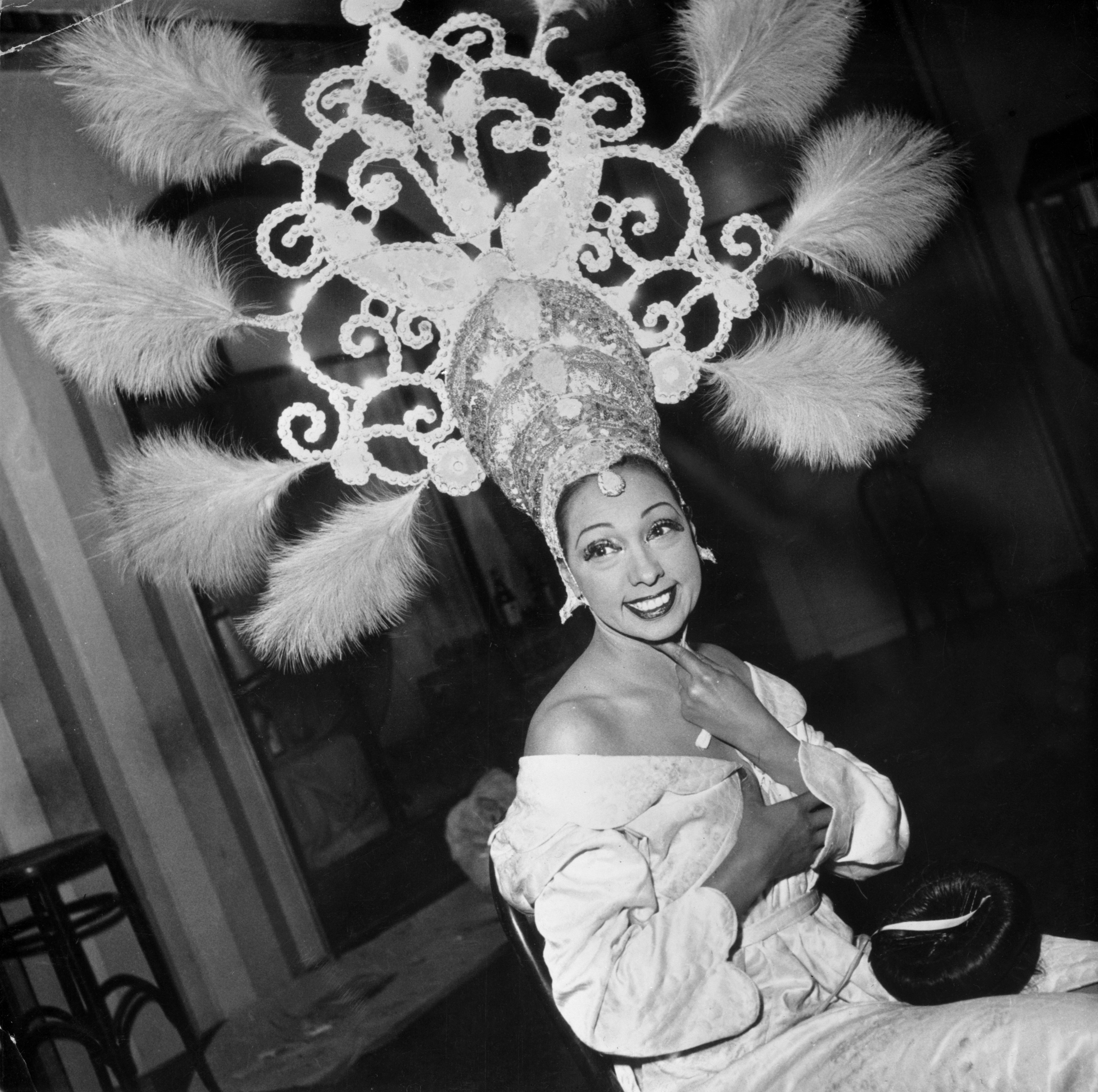 Person wearing an elaborate feathered headdress and elegant off-the-shoulder outfit, smiling toward the camera