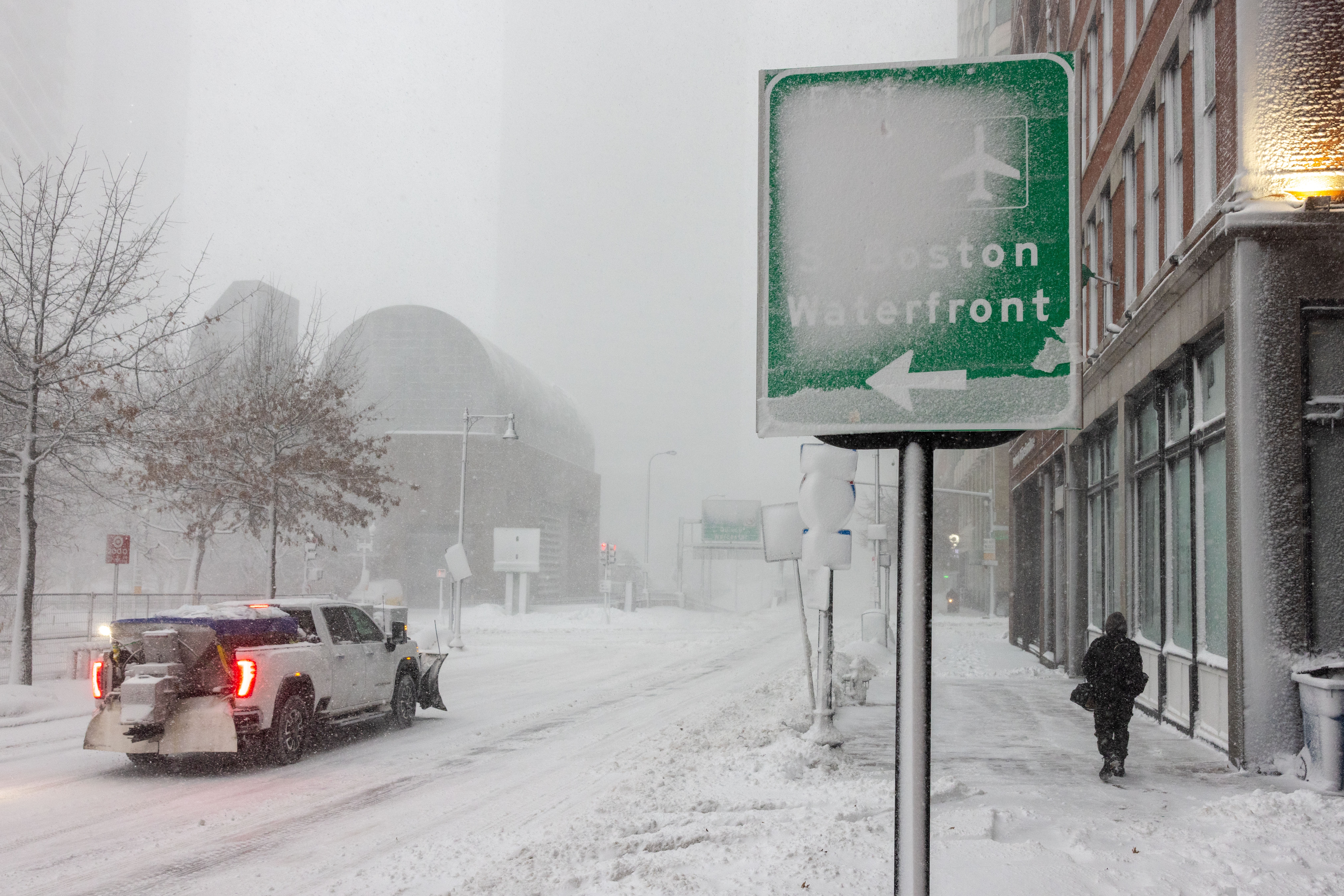 Snødekt gate med en lastebil som brøyter snø og en person som går. Et gateskilt peker mot Boston Waterfront, knapt synlig under snø