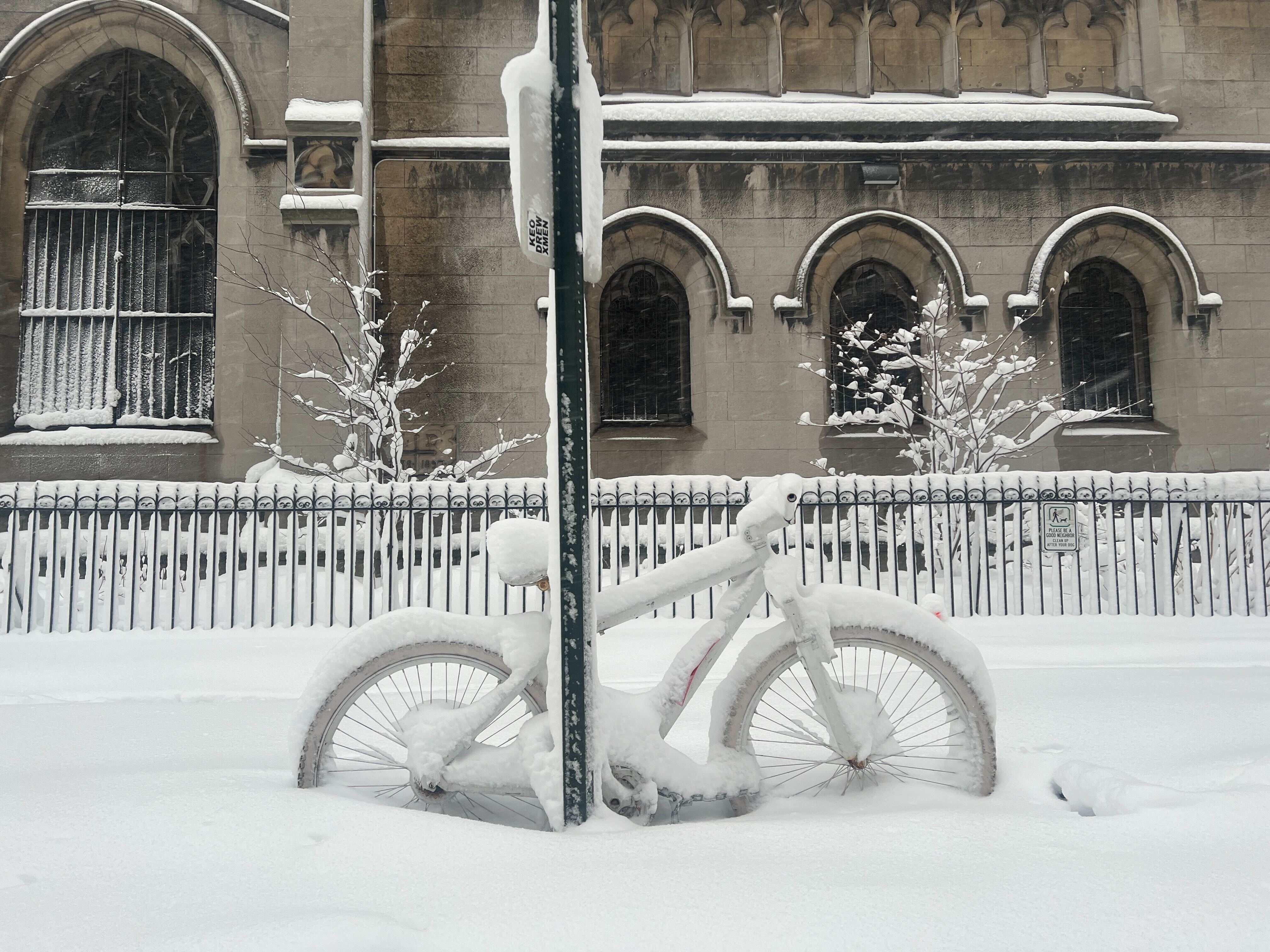 Sykkel dekket av snø er låst til en stolpe foran en bygning med buede vinduer, som viser frem en vinterscene