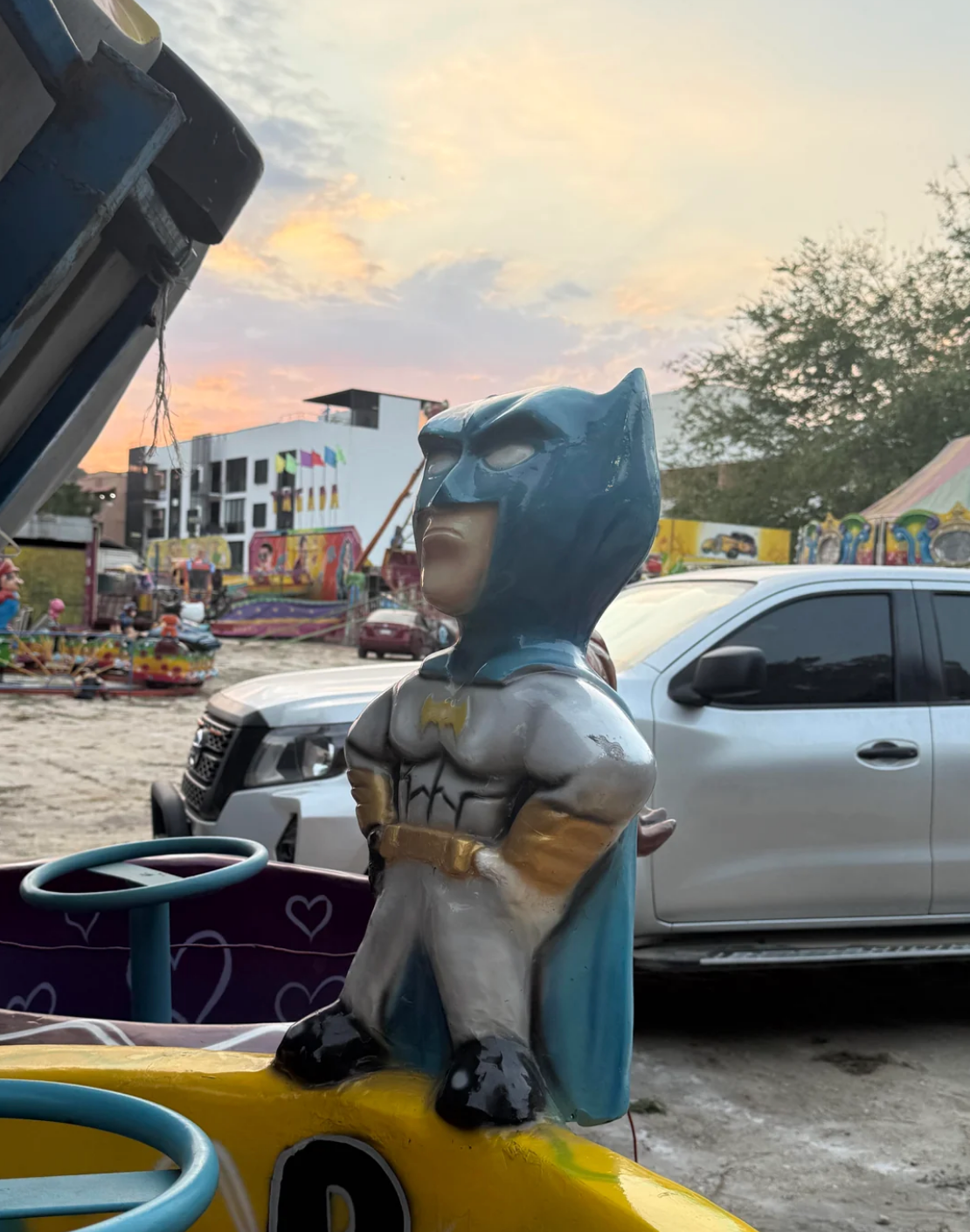 Small Batman statue on a carnival ride seat, with parked cars and tents in the background