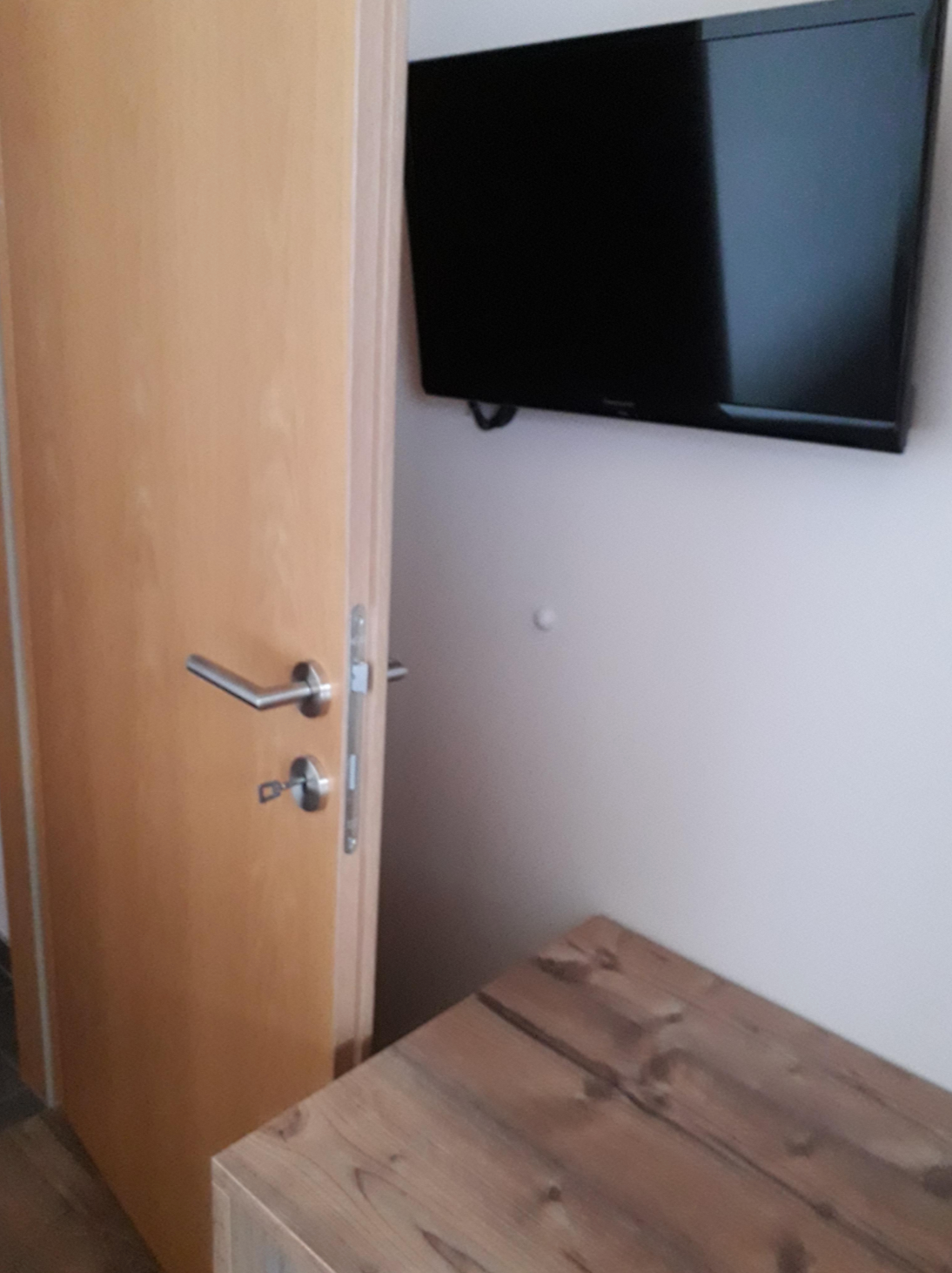 An open wooden door partially obstructs a wall-mounted TV, with a wooden table in the foreground