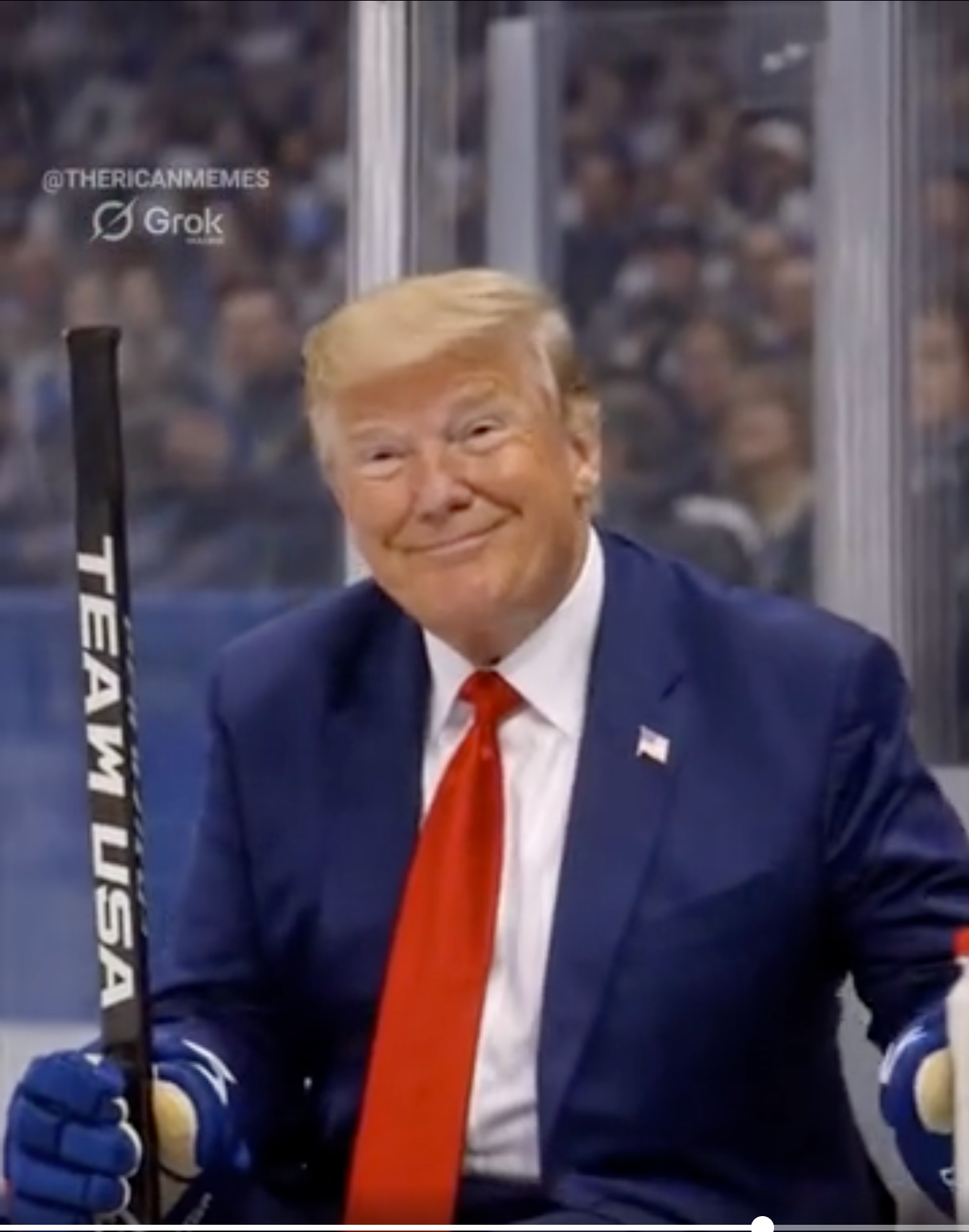 En mann i dress sitter i en hockeyutvisningsboks, holder en hockeykølle og smiler til kameraet