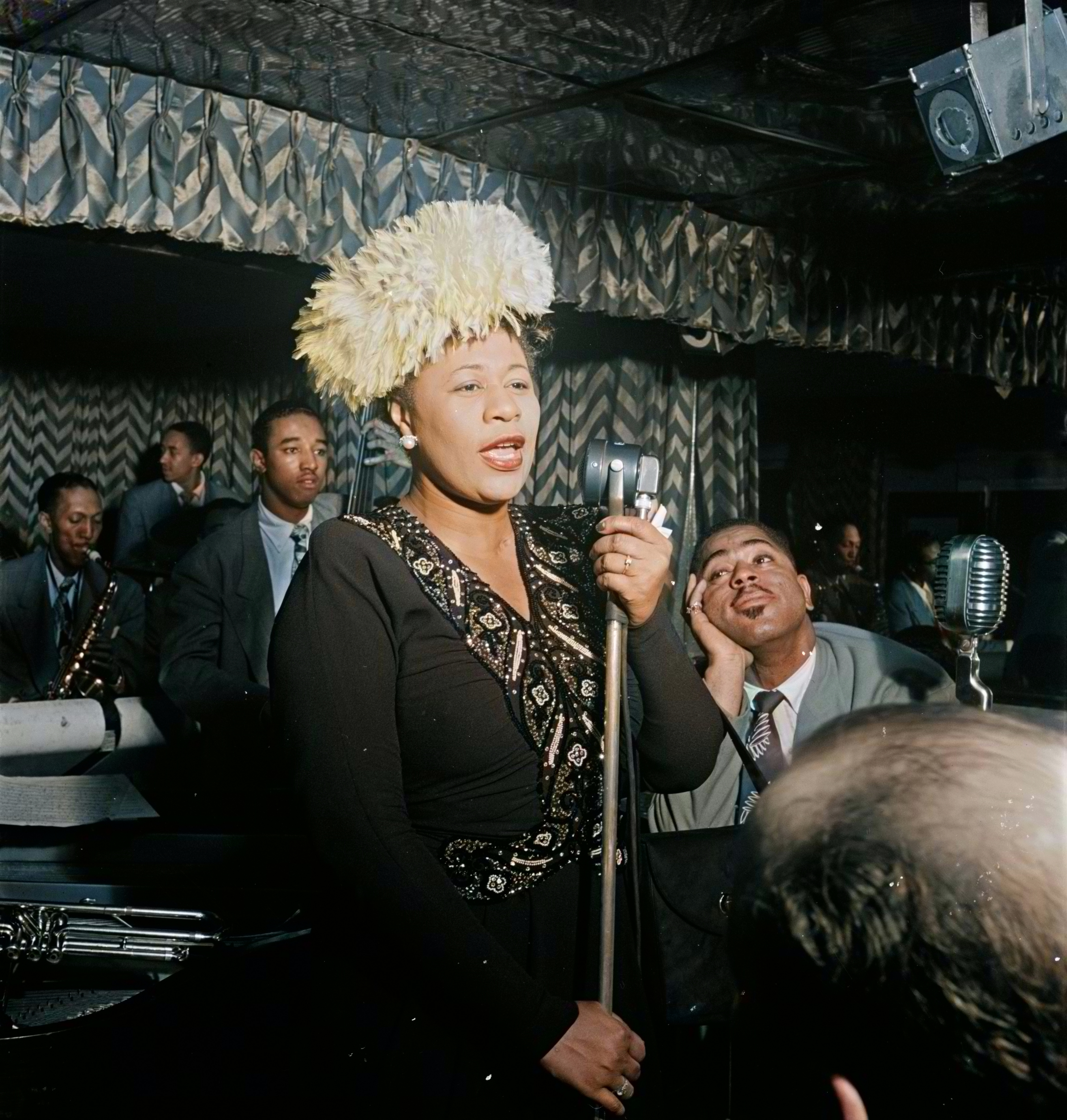 A pistillate   sings into a microphone connected  signifier    with a jazz band, wearing an elaborately decorated achromatic  formal  and a textured hat