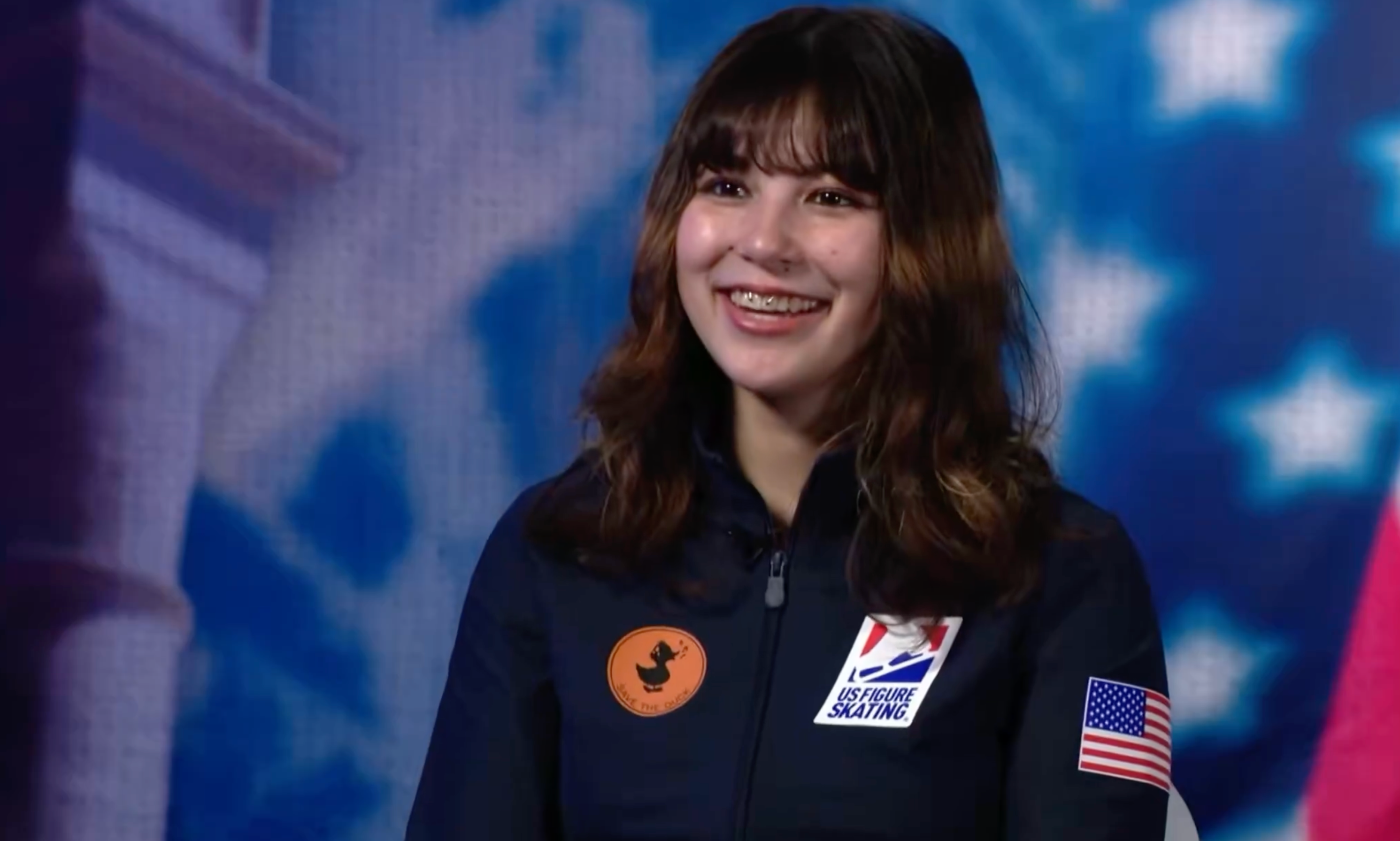 Person med langt hår smiler under et intervju, iført en jakke med et amerikansk flagg og kunstløpslogo