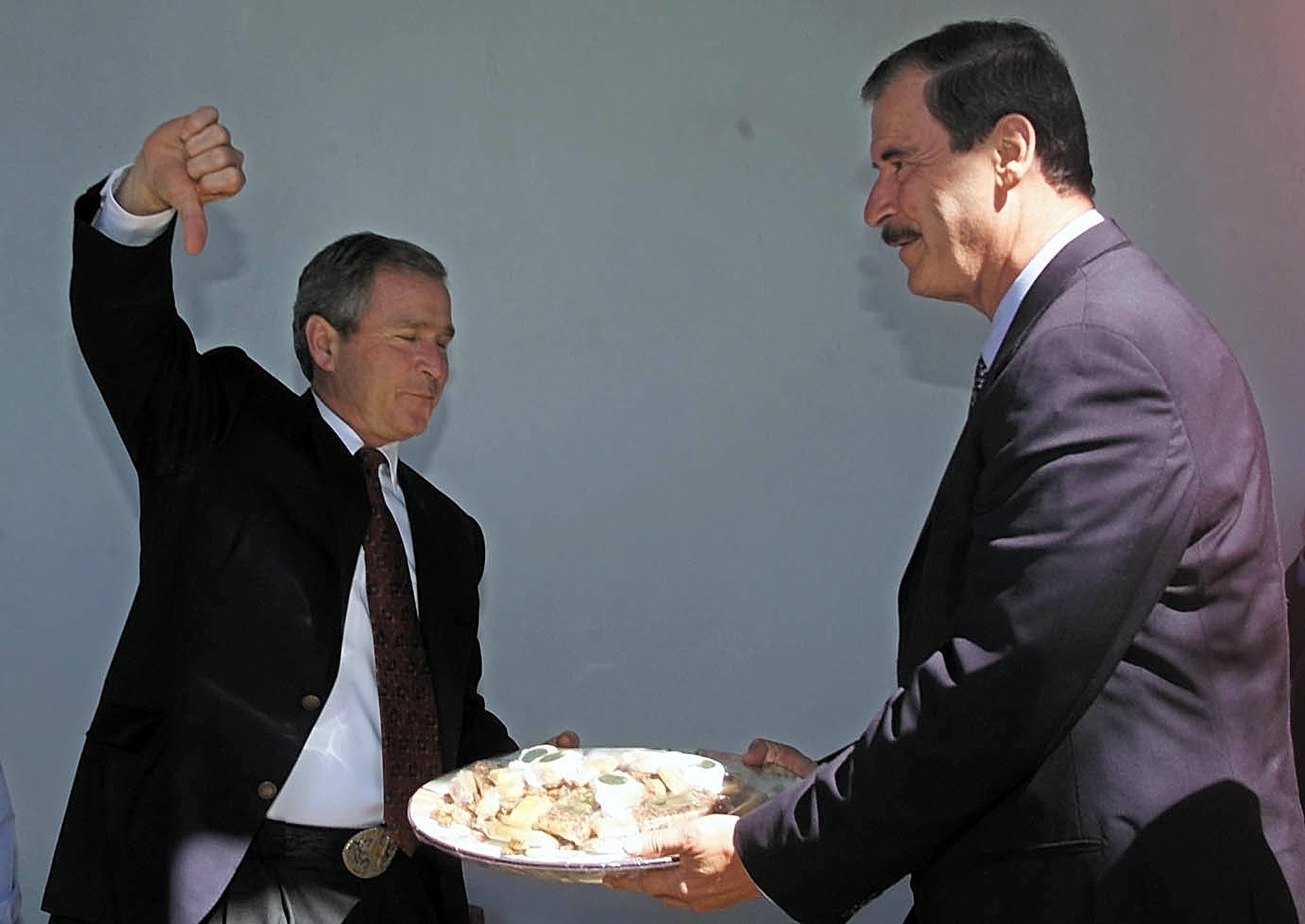 Vicente Fox holder frem en tallerken med mat mens George W. Bush står overfor ham, med høyre arm hevet og gir tommel ned