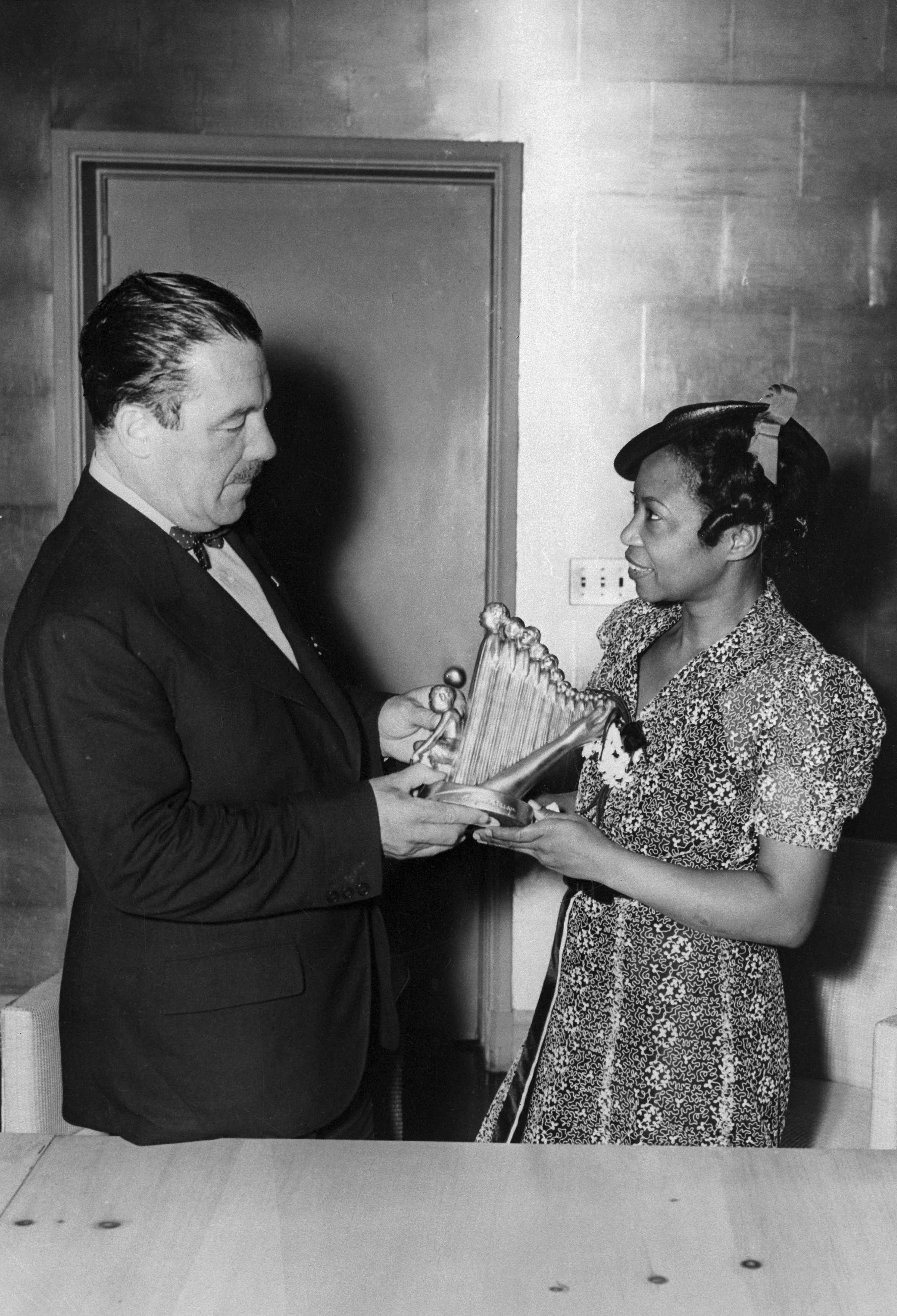 To personer står sammen; en mann i dress deler ut en pris til en kvinne iført mønstret kjole og hatt