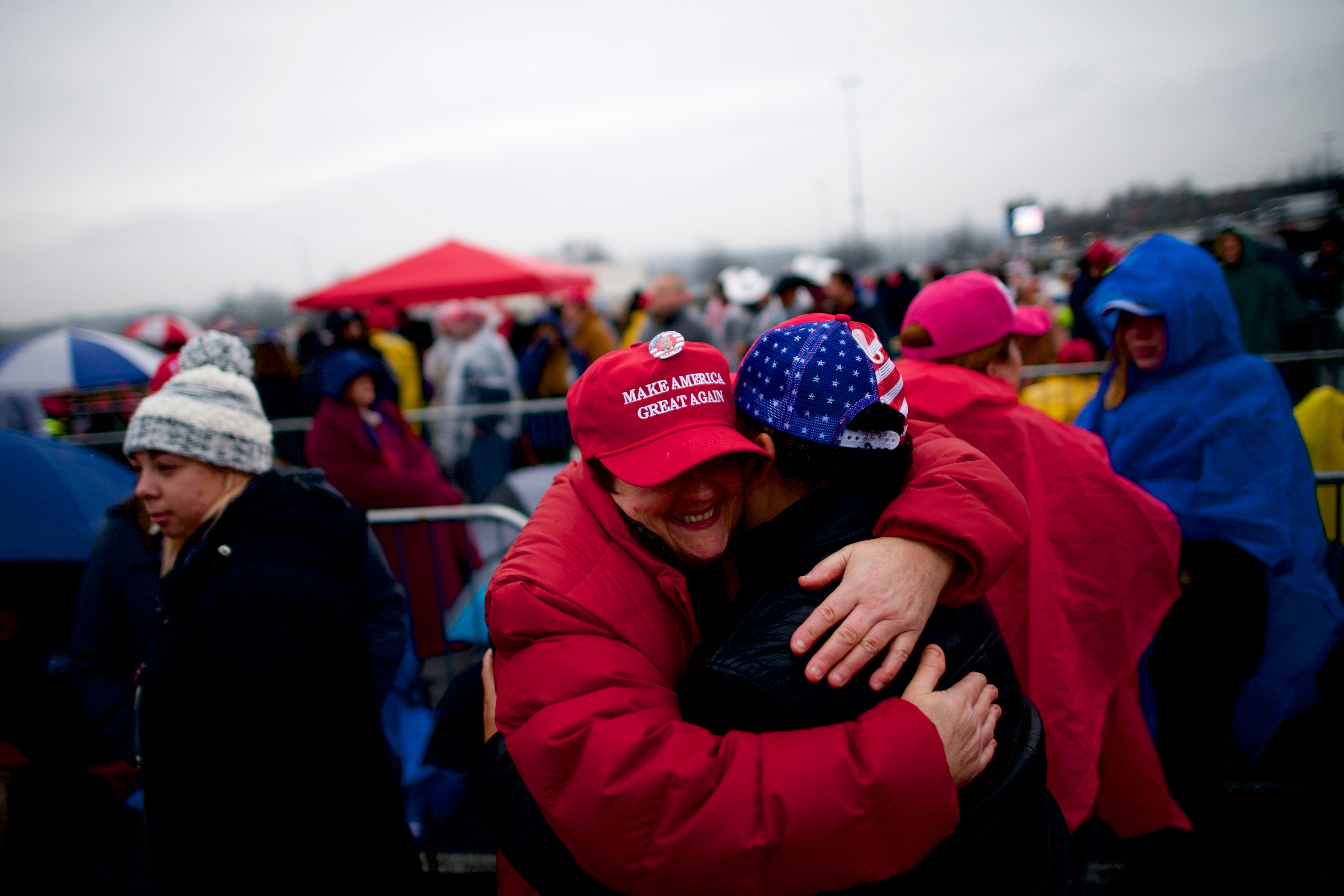 To personer i klær i kaldt vær klemmer på et utendørsarrangement. Man har på seg en "Make America Great Again" hatt. Andre med hatter og regnfrakker er i bakgrunnen