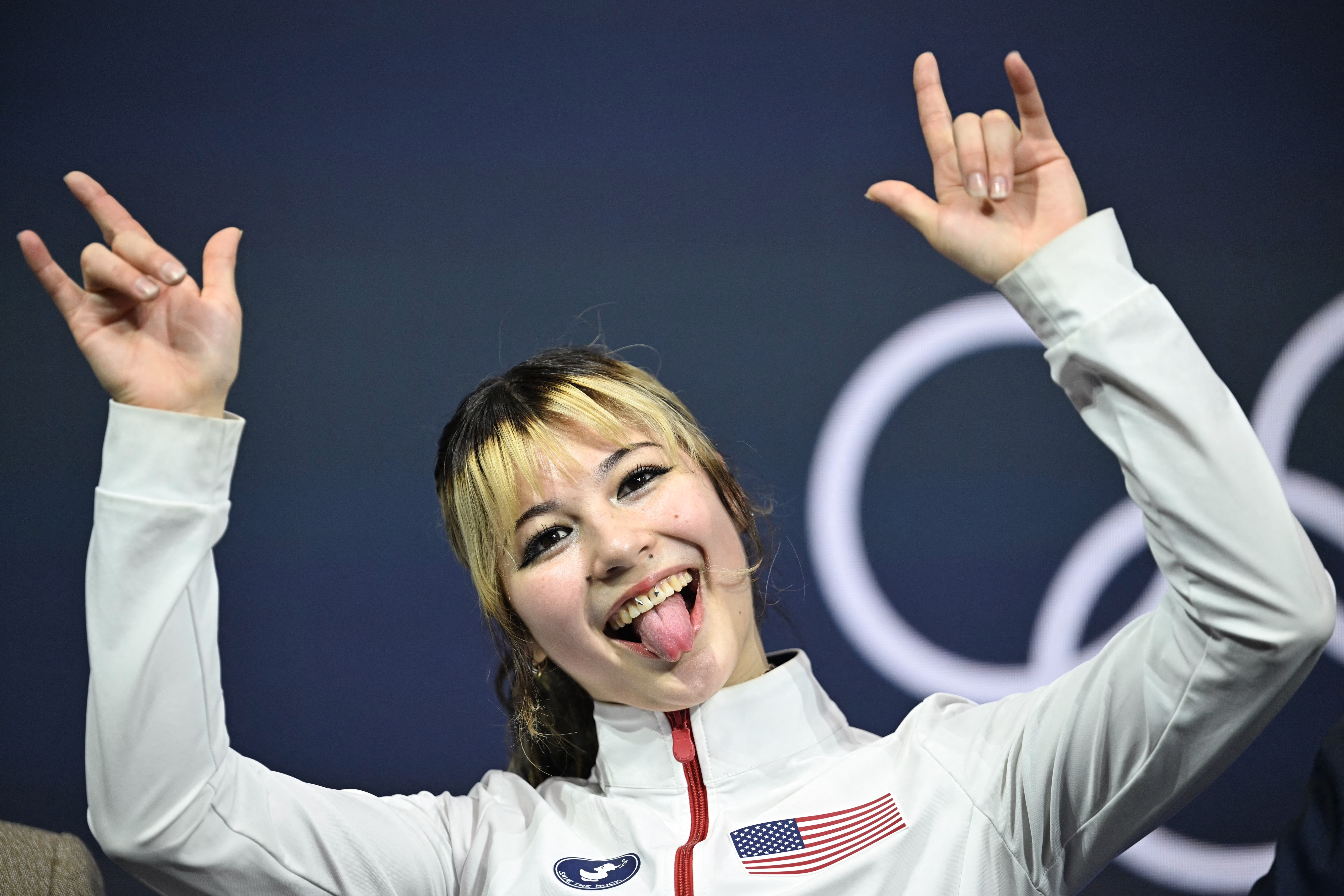 Person successful a achromatic overgarment with a USA flag, smiling with lingua retired and throwing manus signs, lasting successful beforehand of Olympic rings