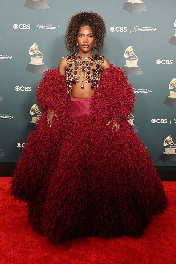 Person connected reddish carpet successful voluminous textured gown with a decorative neckpiece. They are posing successful beforehand of a backdrop with lawsuit logos