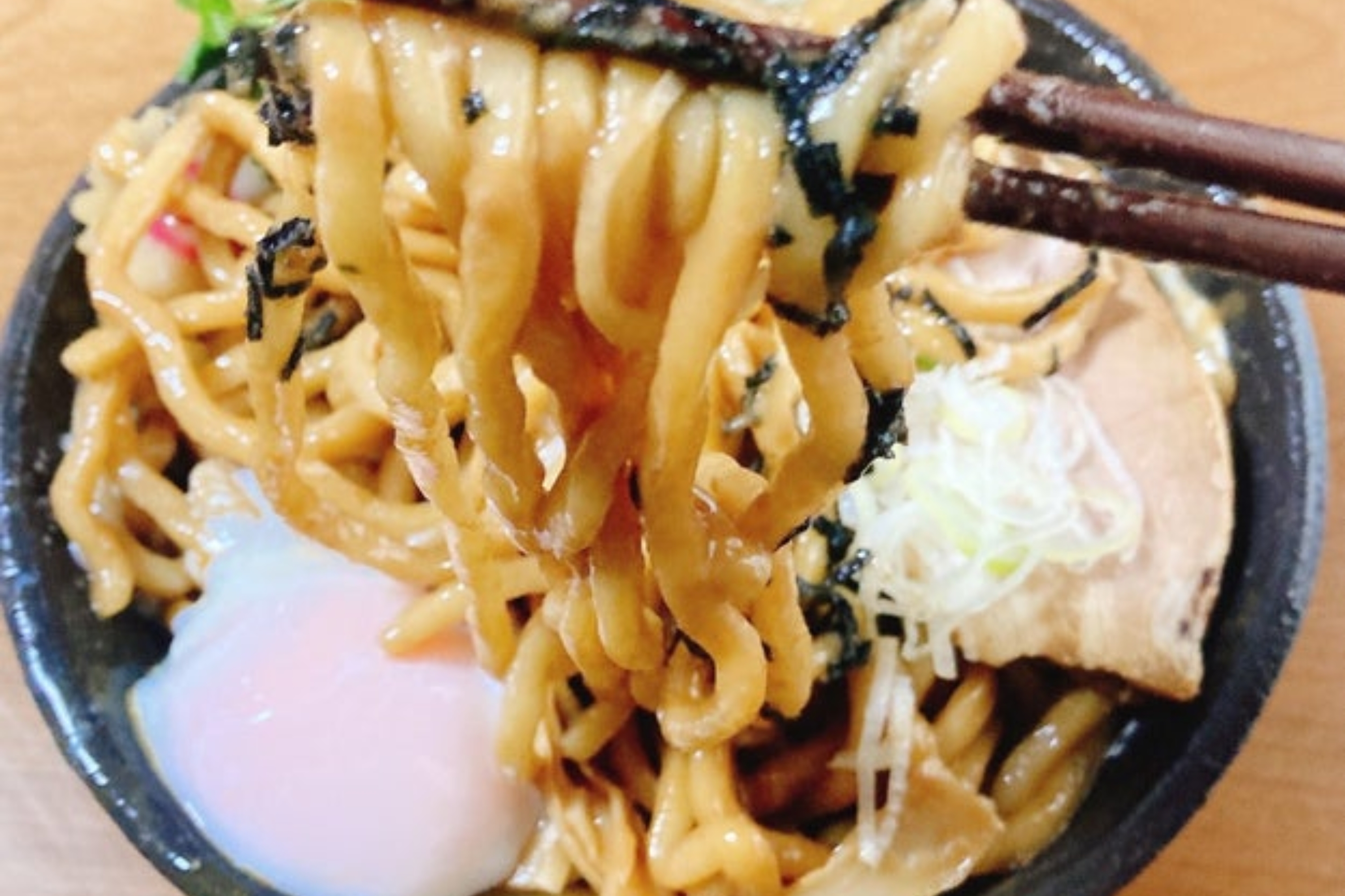 武蔵野アブラ學会の「油そば」。麺を箸で持ち上げているところ。麺のうえには、半熟卵・ネギ・焼豚がトッピングされていて美味しそうだ。