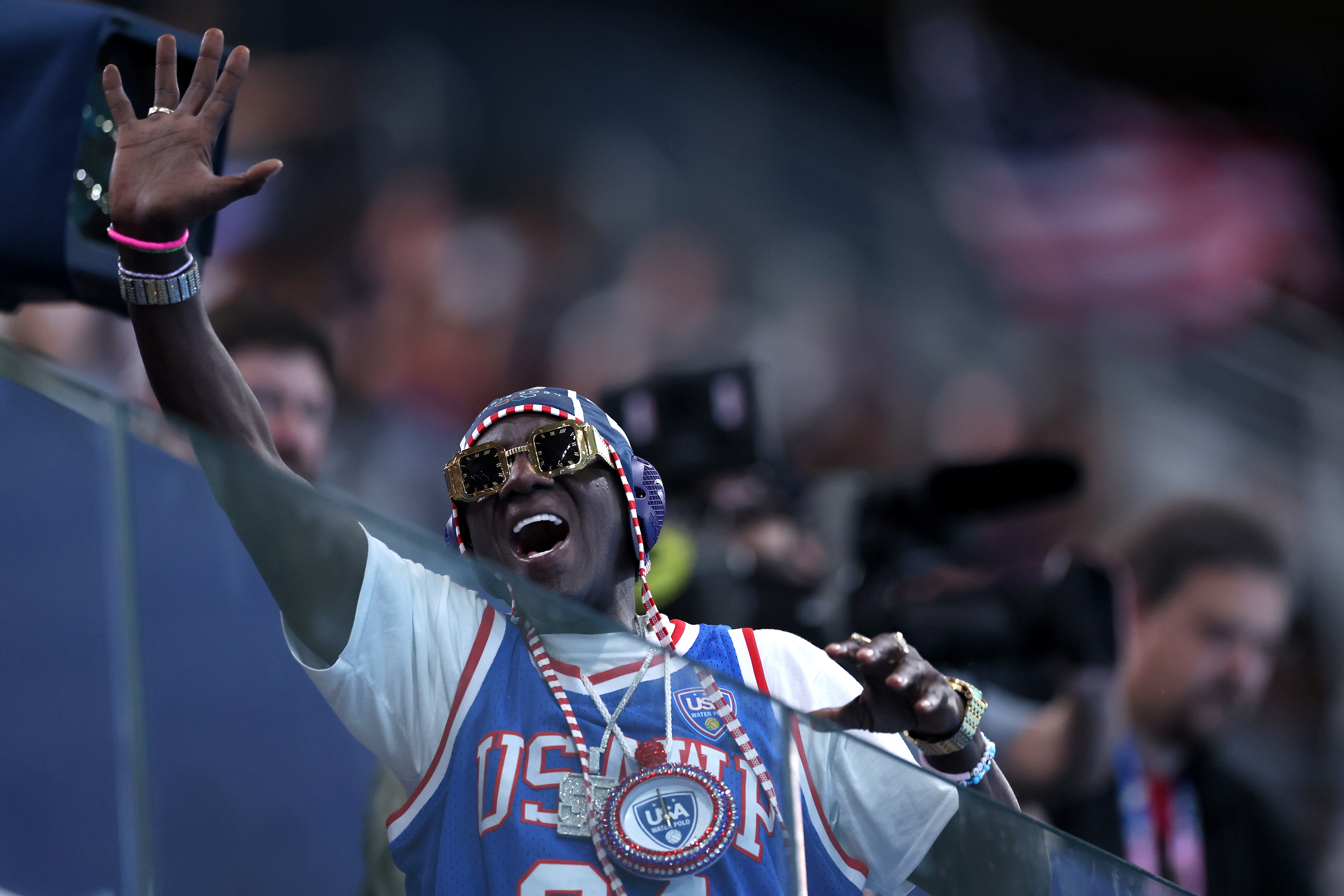 Person i sporty antrekk med store solbriller og et stort klokkekjede vinker energisk på et arrangement, omgitt av kameraer