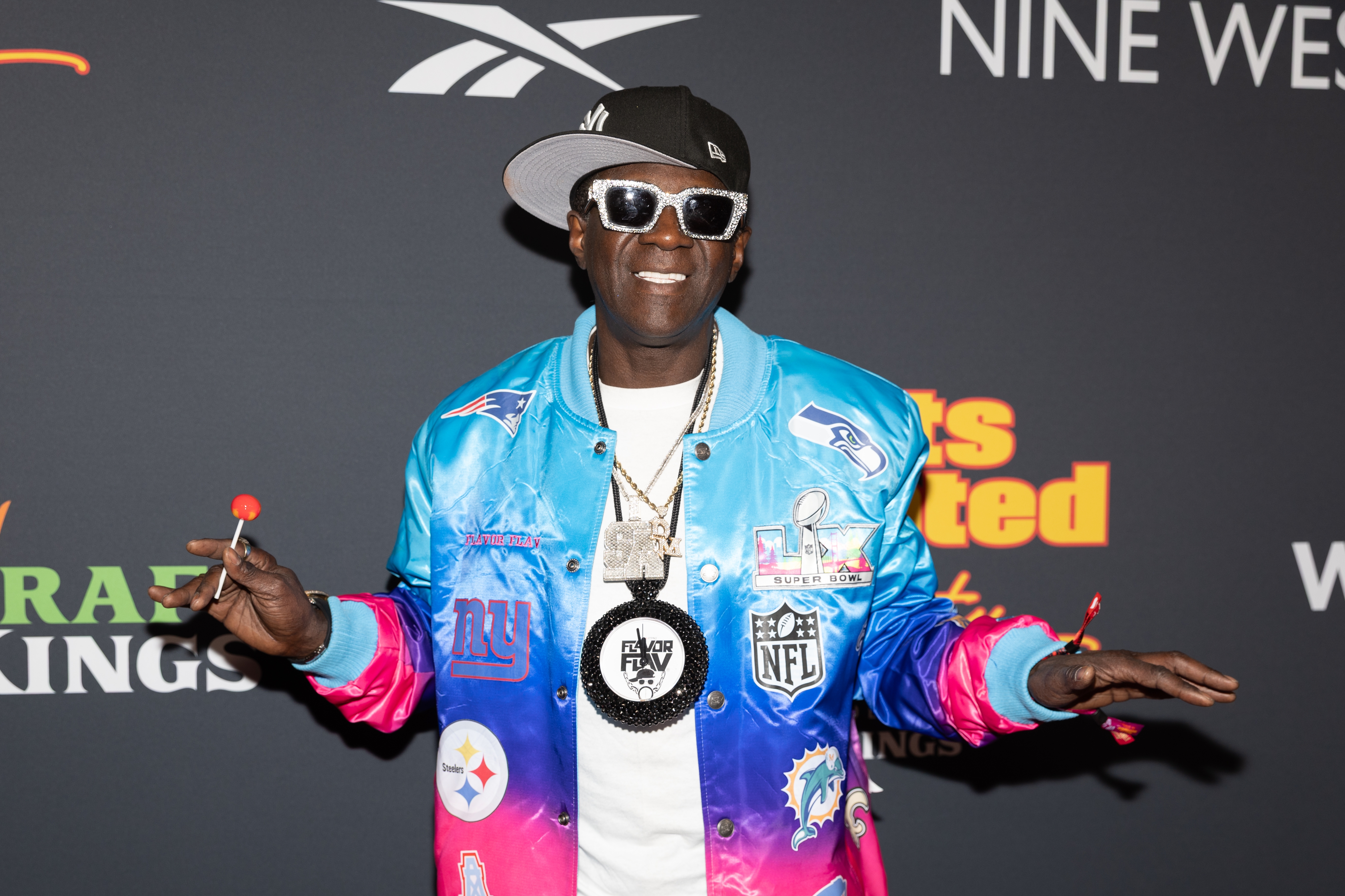 Kjendis iført en livlig jakke med sportstema, overdimensjonert klokkekjede, caps og solbriller på den røde løperen. Holder en slikkepinne