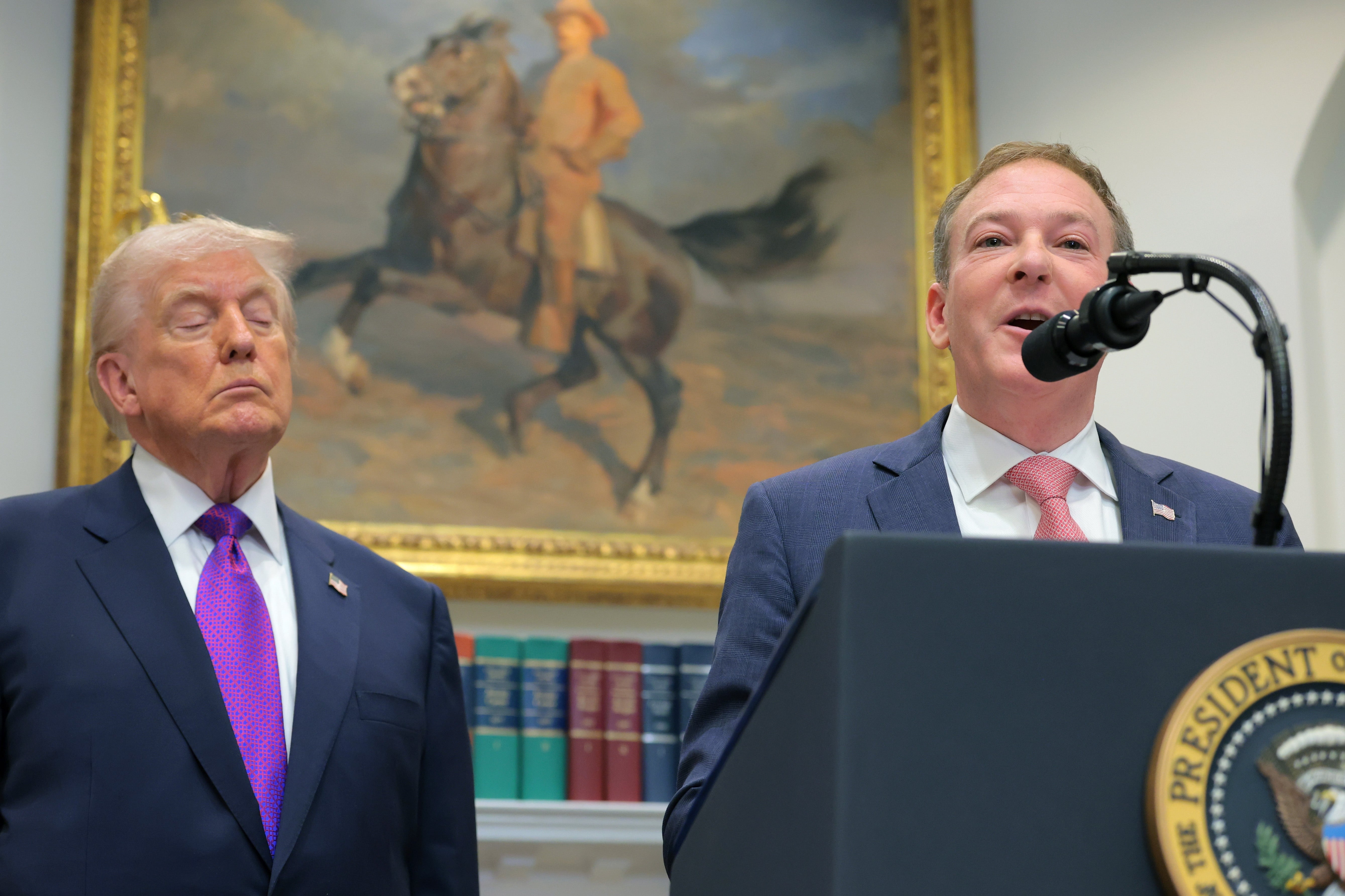 Trump dan Lee Zeldin berada di podium, Zeldin berbicara, dan mata Trump tampak tertutup. Lukisan seorang pria menunggang kuda terlihat di latar belakang