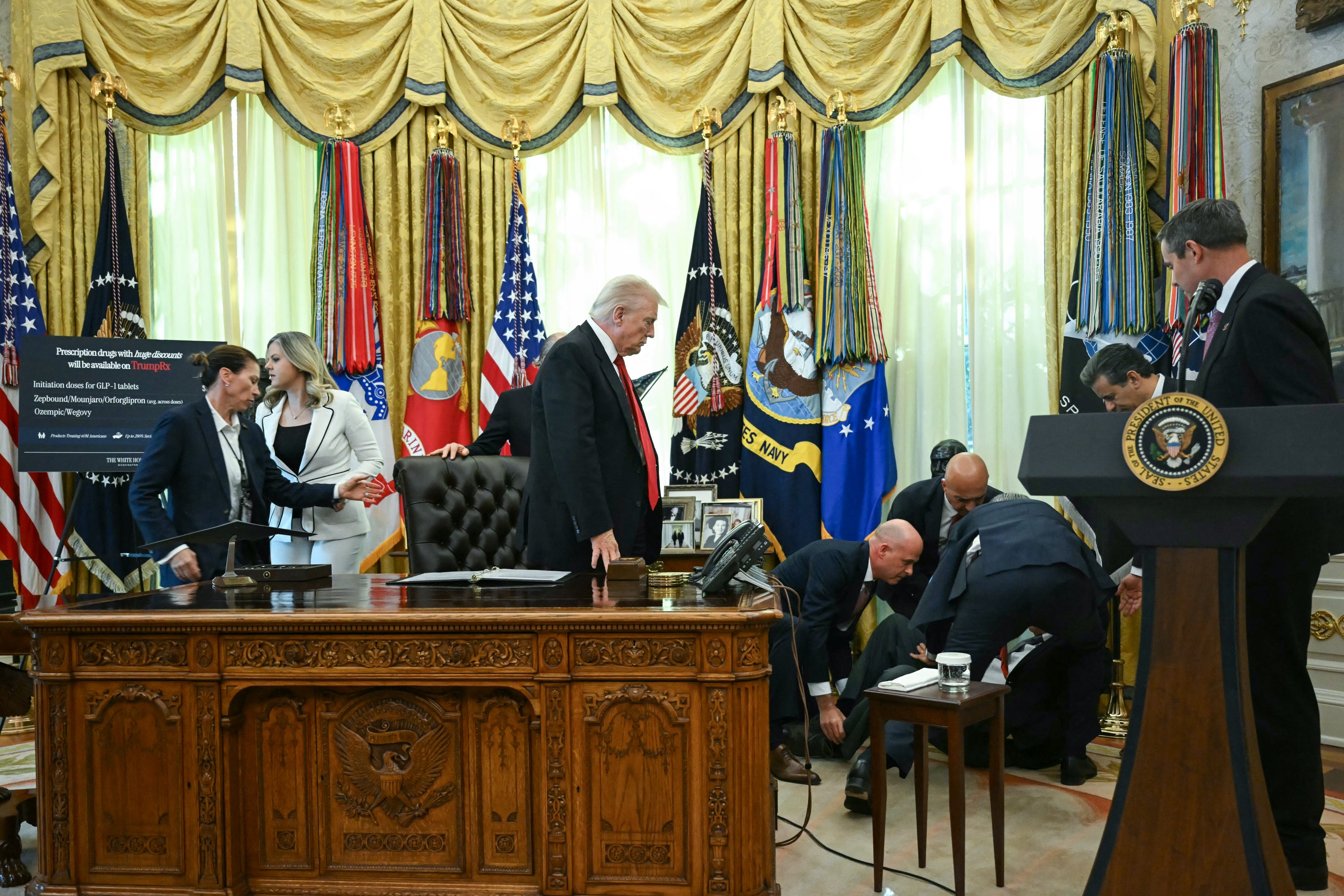 Trump berdiri di belakang meja tegas di Ruang Oval ketika orang-orang di dekatnya merawat orang yang sakit