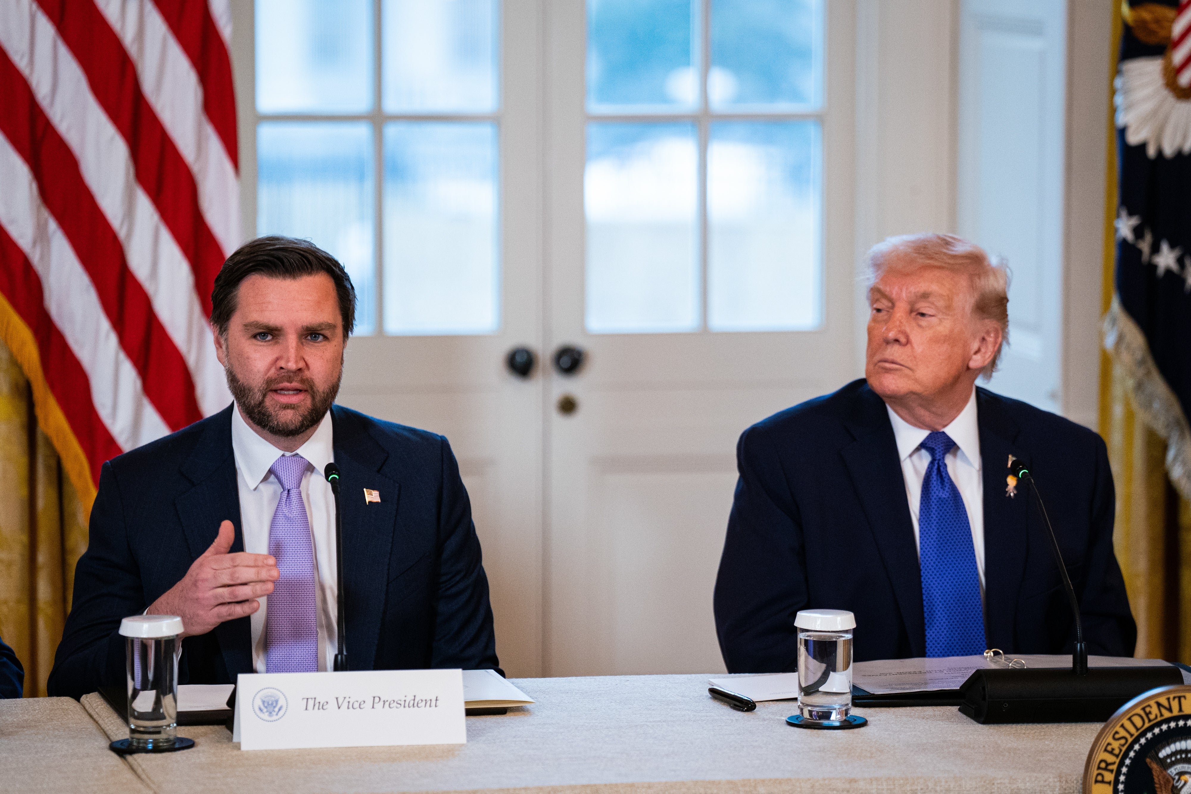 JD Vance dan Trump duduk di meja konferensi dengan mikrofon dan gelas air; Vance berbicara, Trump mendengarkan, tapi dia terlihat sedikit kesal, dalam suasana formal