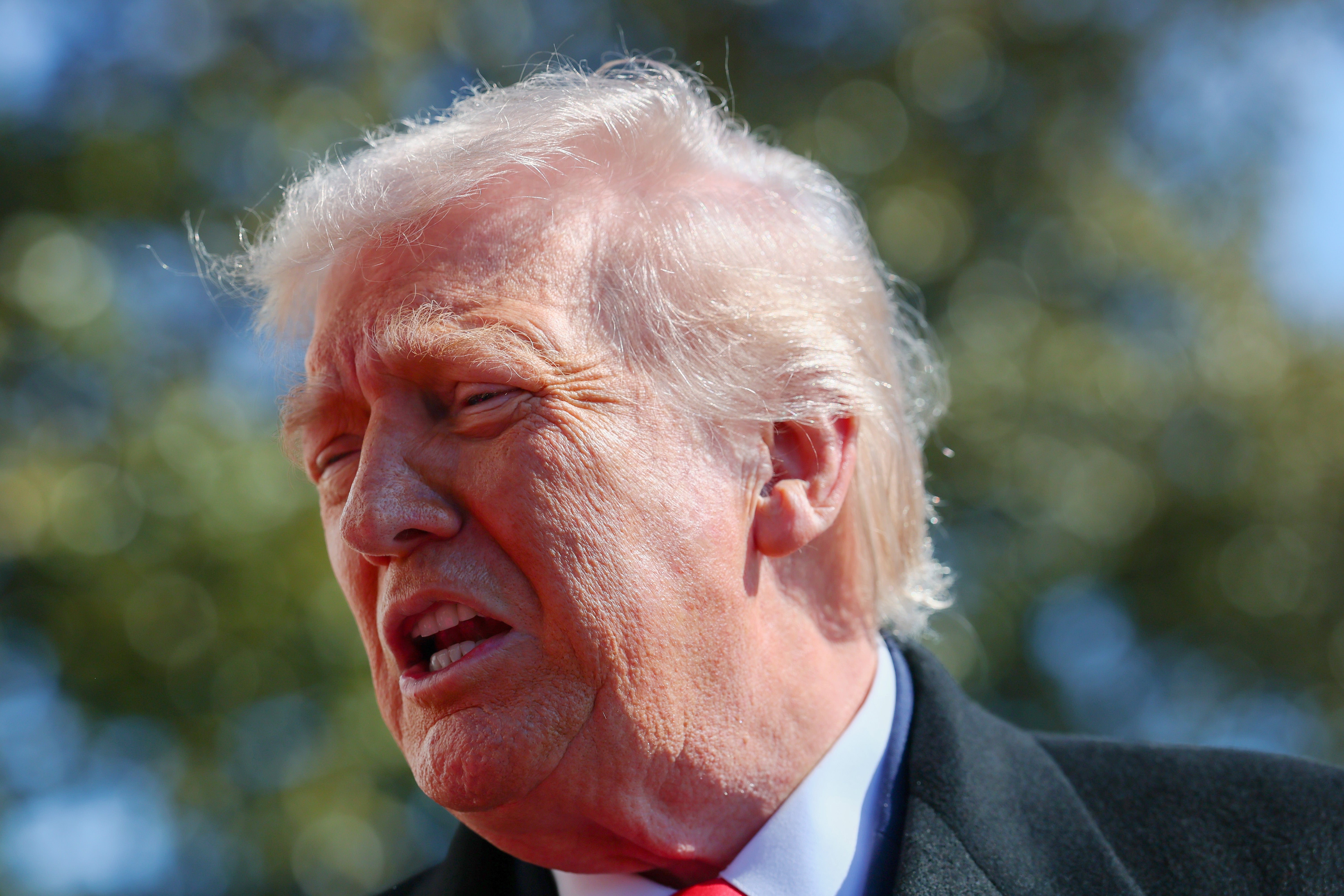 Donald Trump tampak tidak nyaman, menyipitkan mata dalam setelan jas dan berbicara di luar ruangan, latar belakang sedikit kabur
