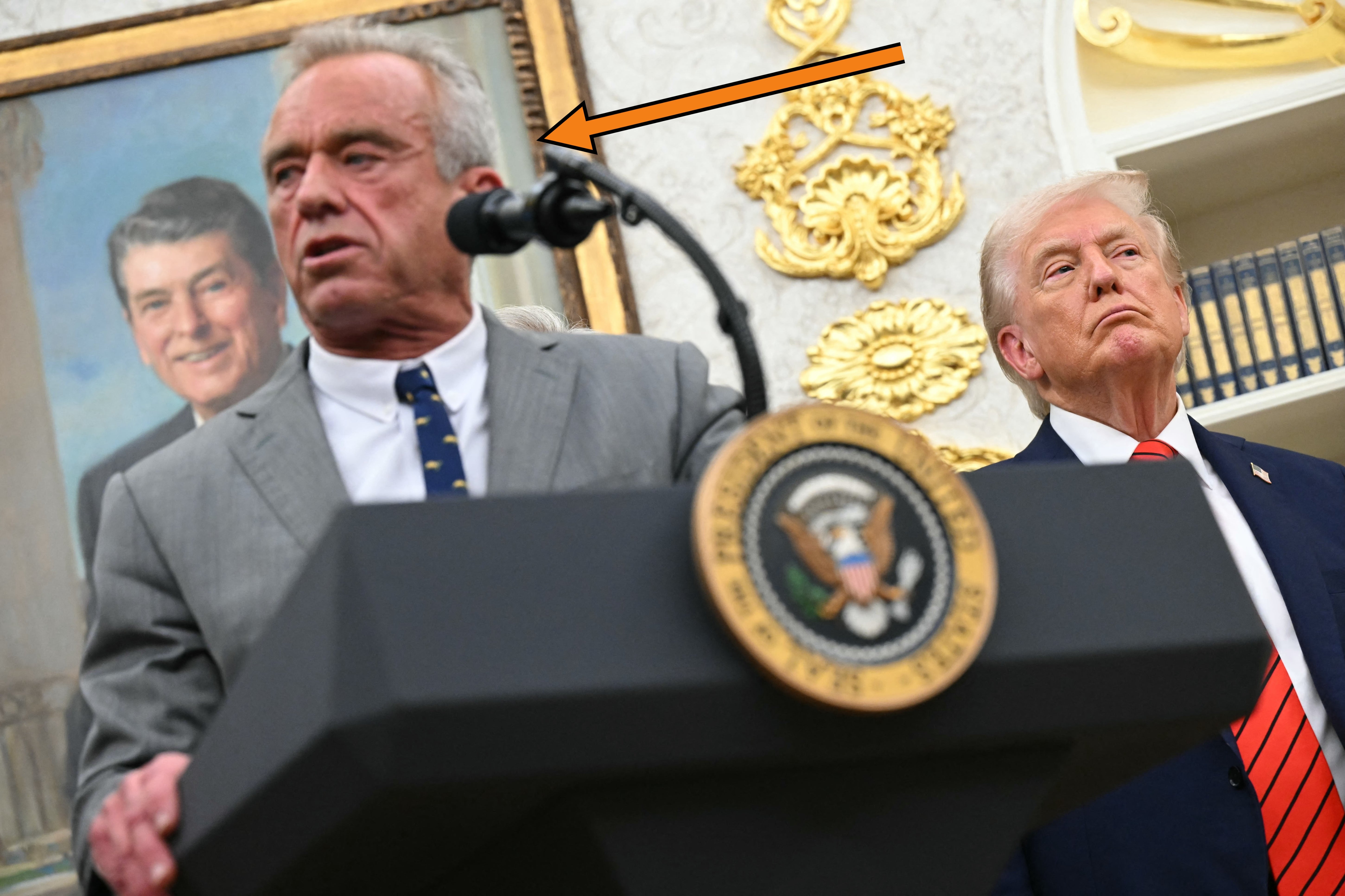 Dua pria di podium; RFK Jr dalam setelan abu-abu berbicara, Trump dalam setelan jas dengan dasi merah, tampak bosan. Potret mantan presiden (Reagan) ada di latar belakang