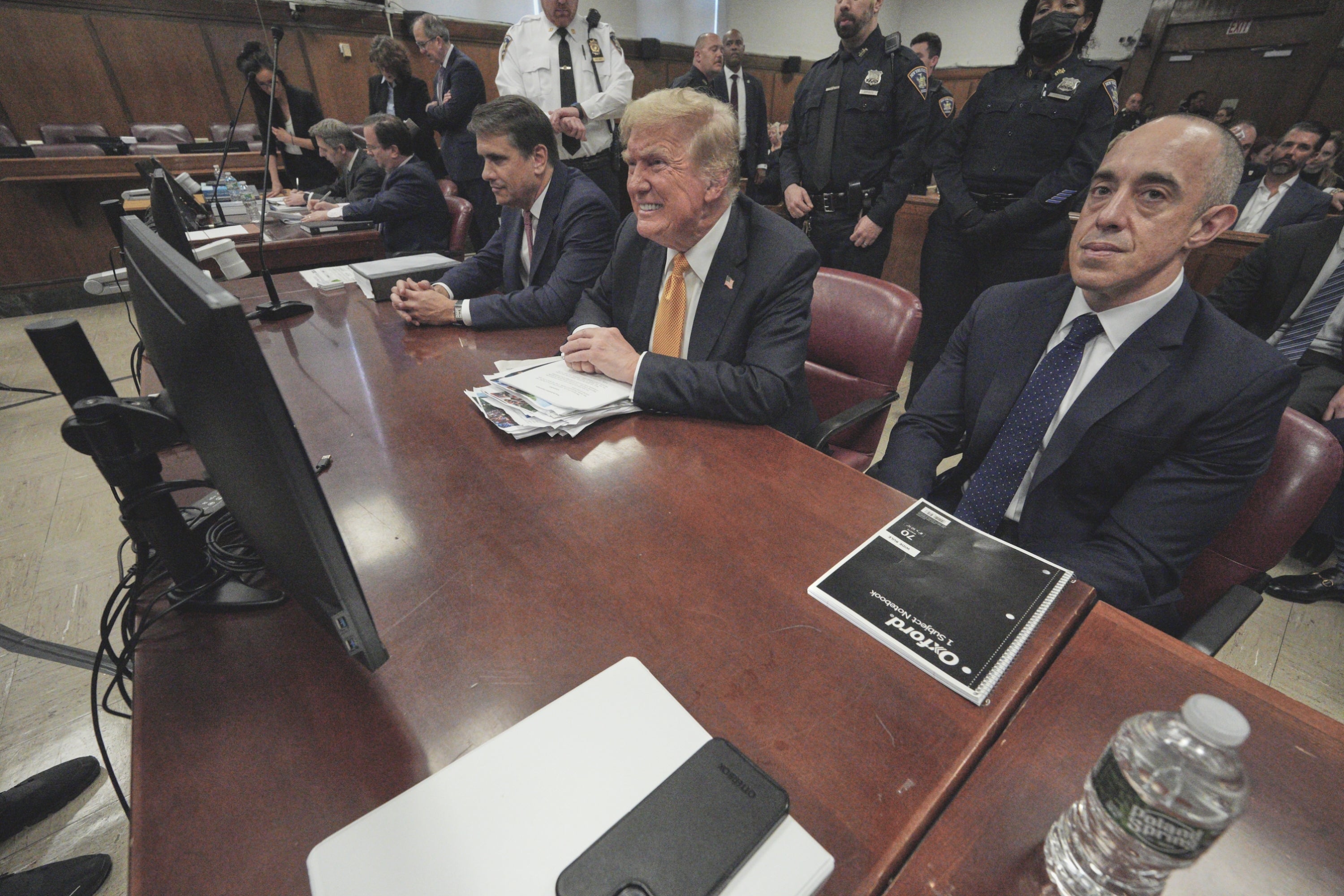 Sekelompok pria berjas duduk di meja ruang sidang, mendengarkan dengan penuh perhatian. Trump berada di tengah-tengah mereka dan berusaha keras. Berbagai pejabat dan personel keamanan terlihat di latar belakang
