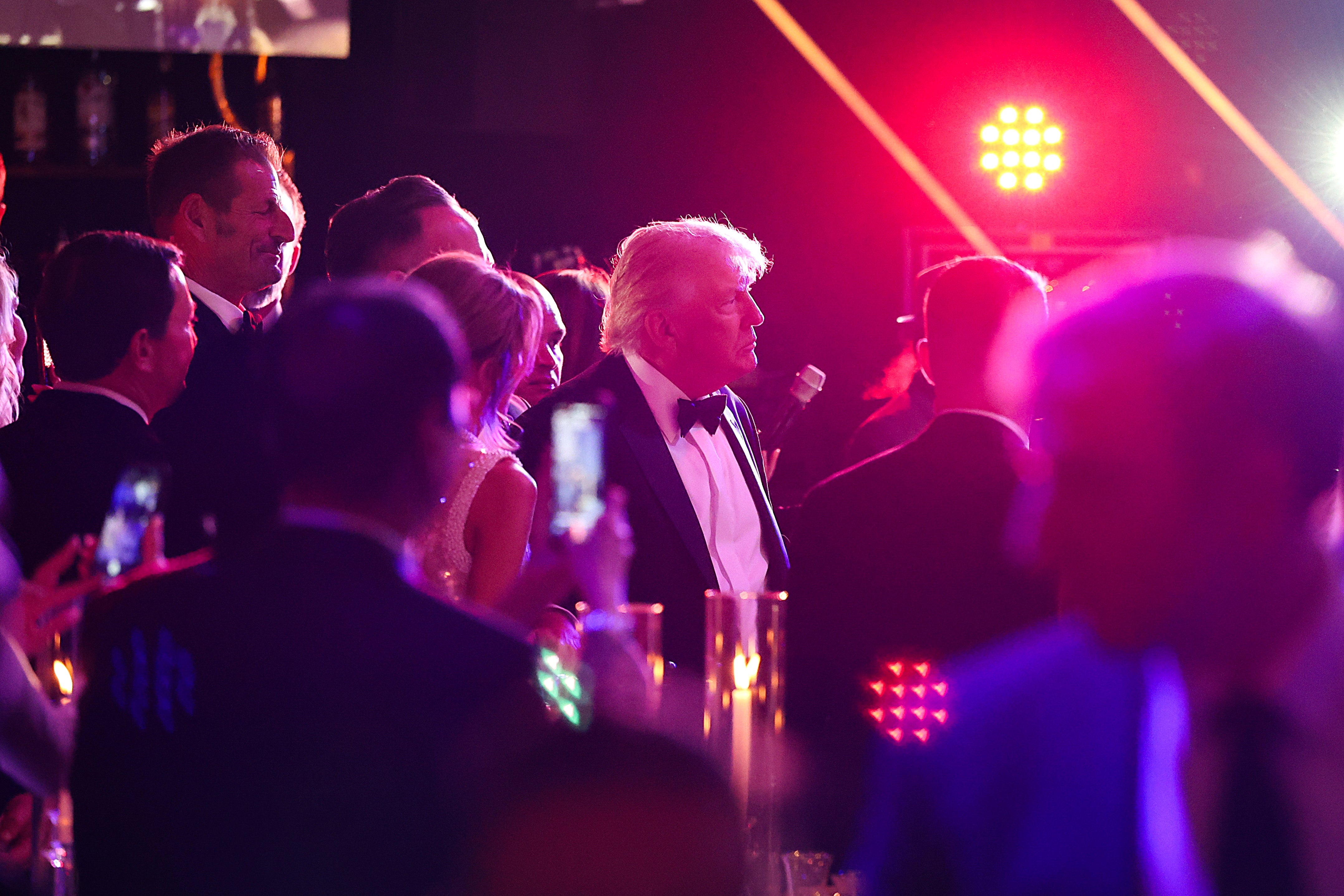 Sekelompok orang, termasuk Trump dengan tuksedo, berkumpul di acara formal dengan lampu terang dan kamera; Trump menatap lurus ke depan