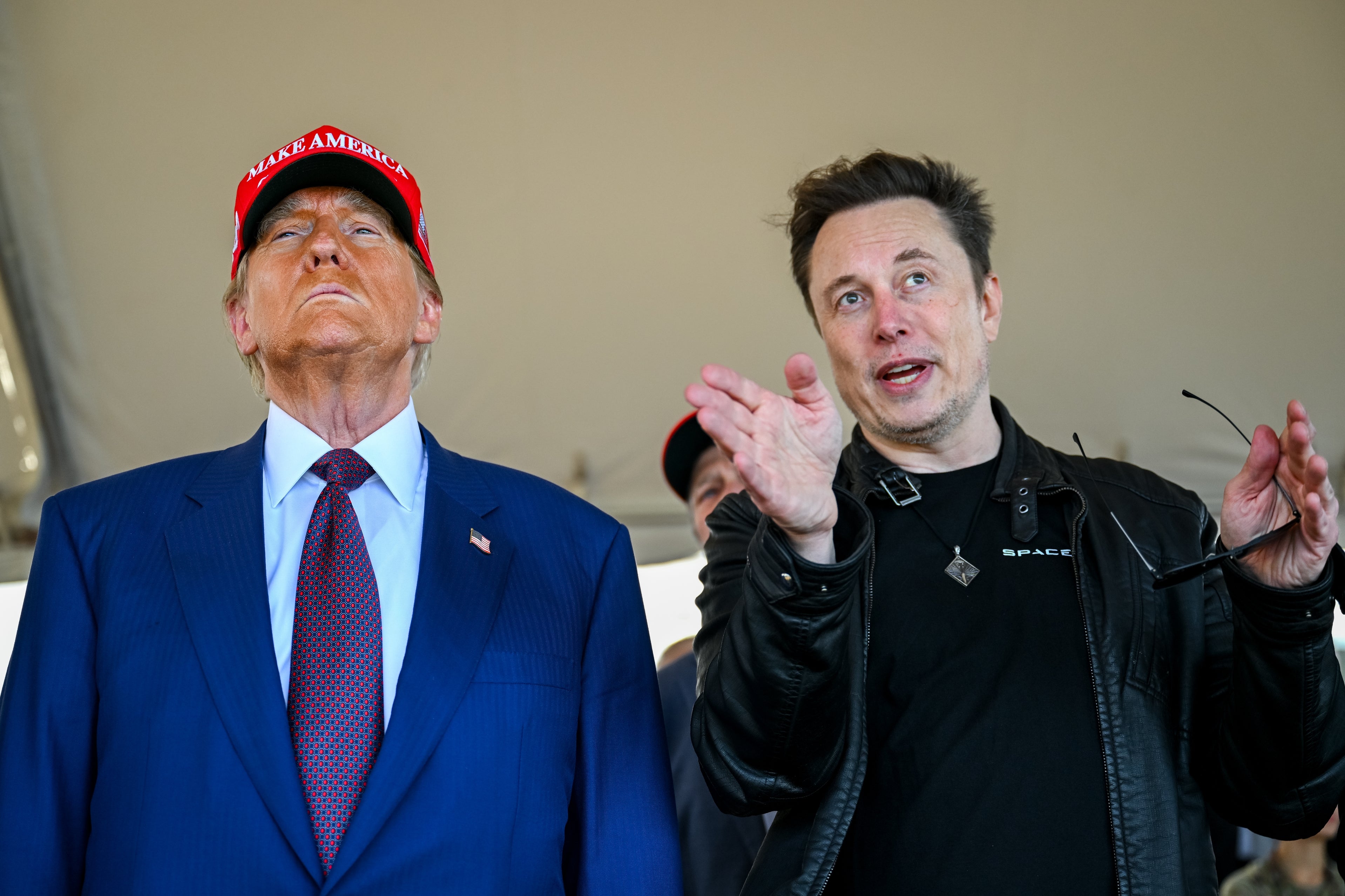Trump berdiri di samping Elon di sebuah acara. Trump mengenakan setelan jas, Elon mengenakan kaos oblong dan jaket kulit; Elon memberi isyarat ke arah sesuatu dengan mulut terbuka, dan Trump berdiri dengan tenang mengenakan topi merah
