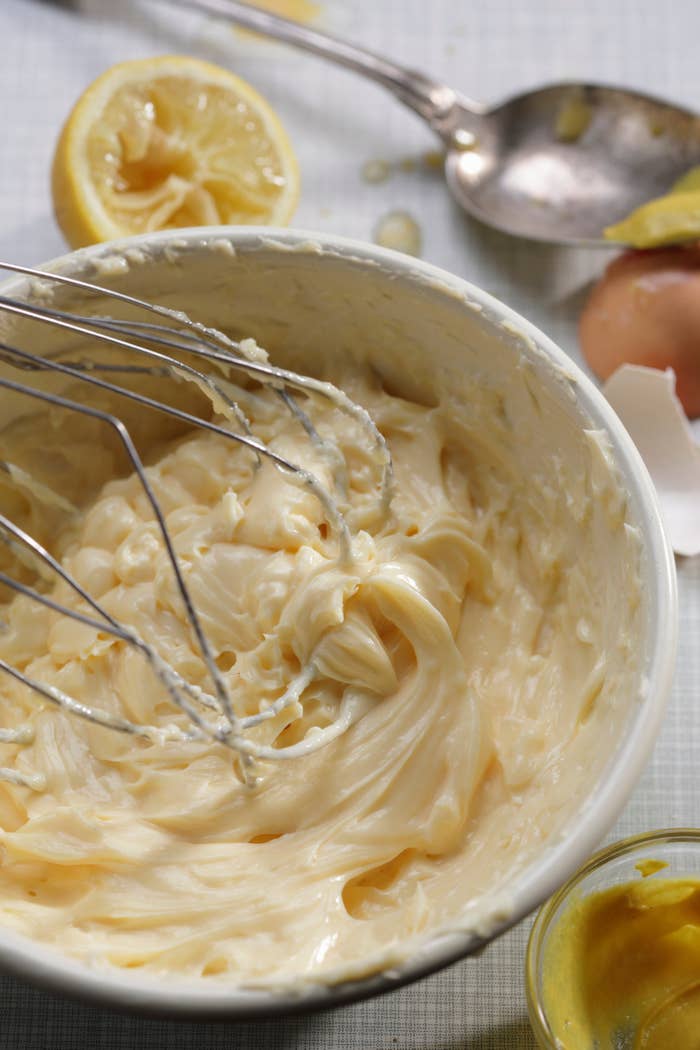 Bowl of homemade mayonnaise with a whisk, adjacent  to a cracked egg, citrus  half, and a spoon