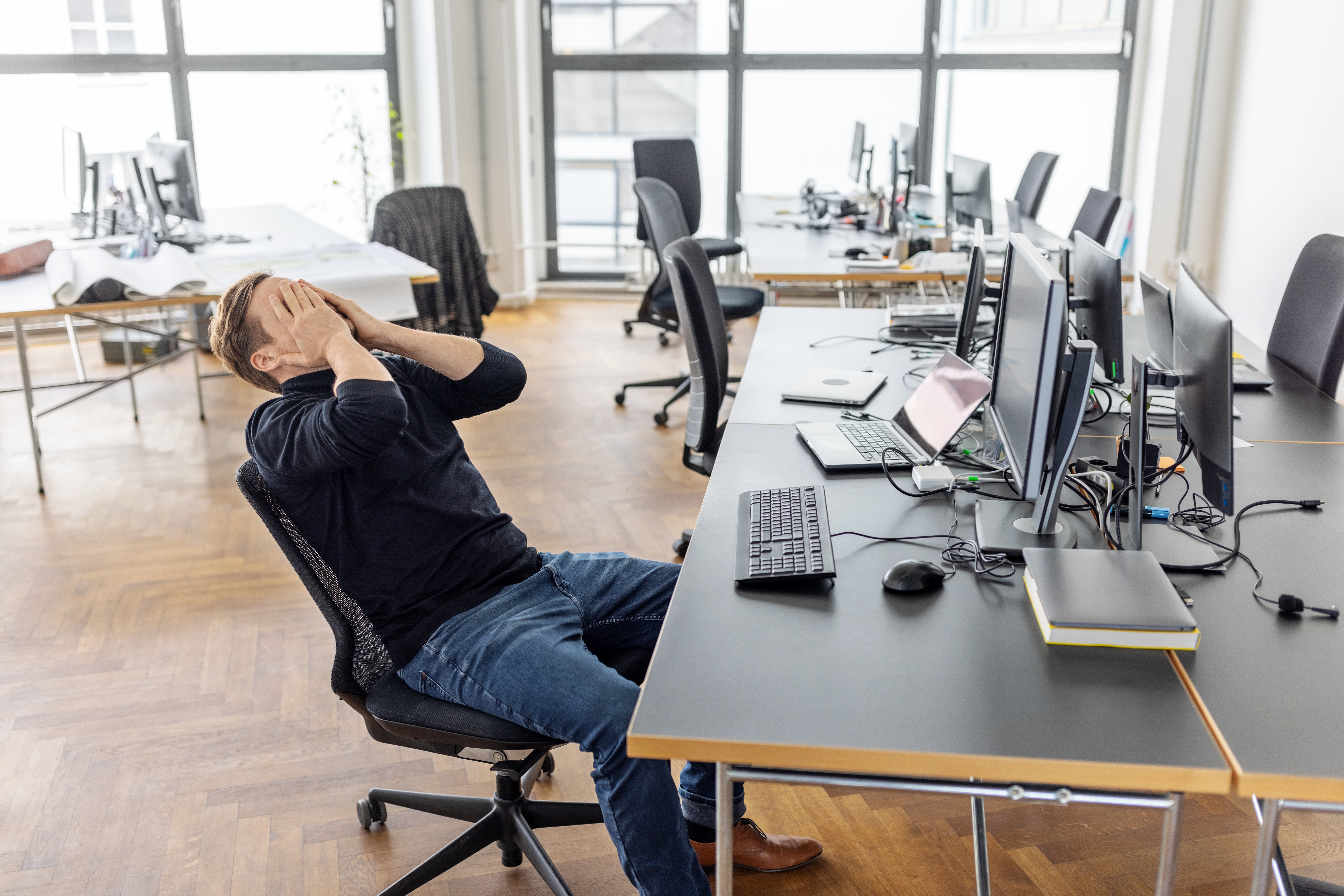 Man successful an bureau sitting astatine a desk, appearing frustrated with his caput successful his hands, surrounded by machine monitors