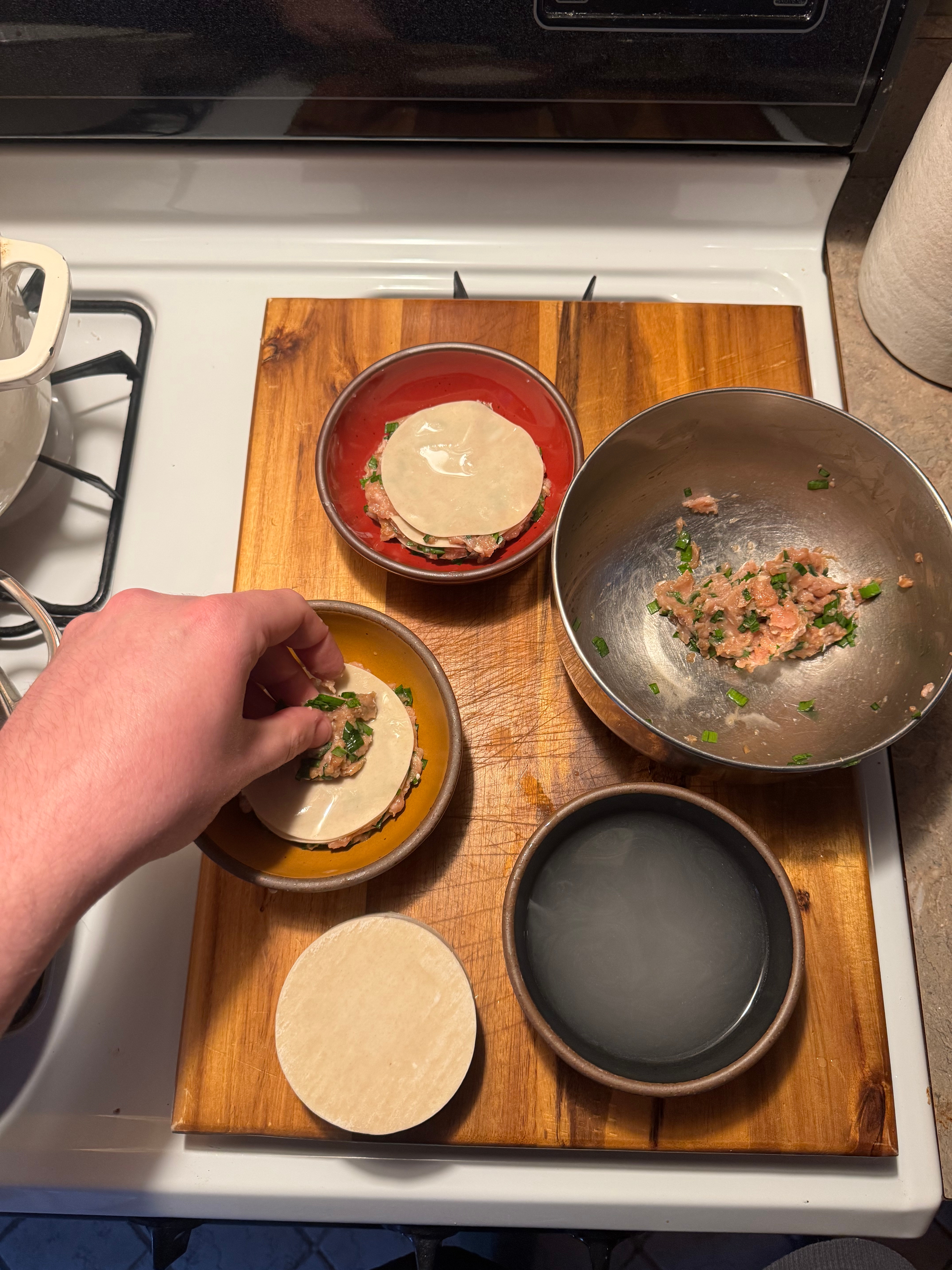 A person prepares dumplings on a wooden board with various fillings and wrappers, using different bowls for ingredients