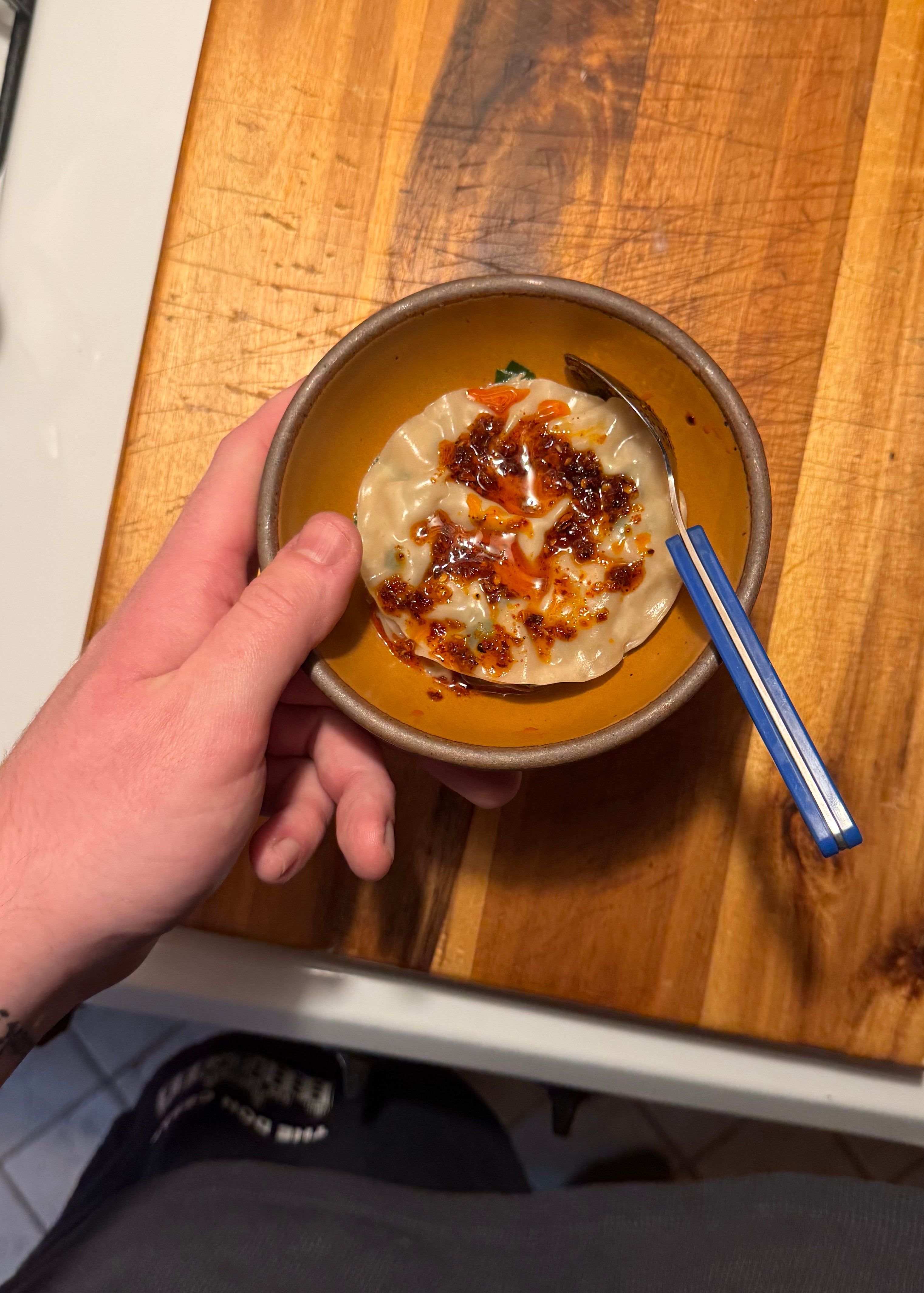 A person holds a bowl of dumplings topped with chili oil and herbs on a wooden board, with part of a stove visible nearby