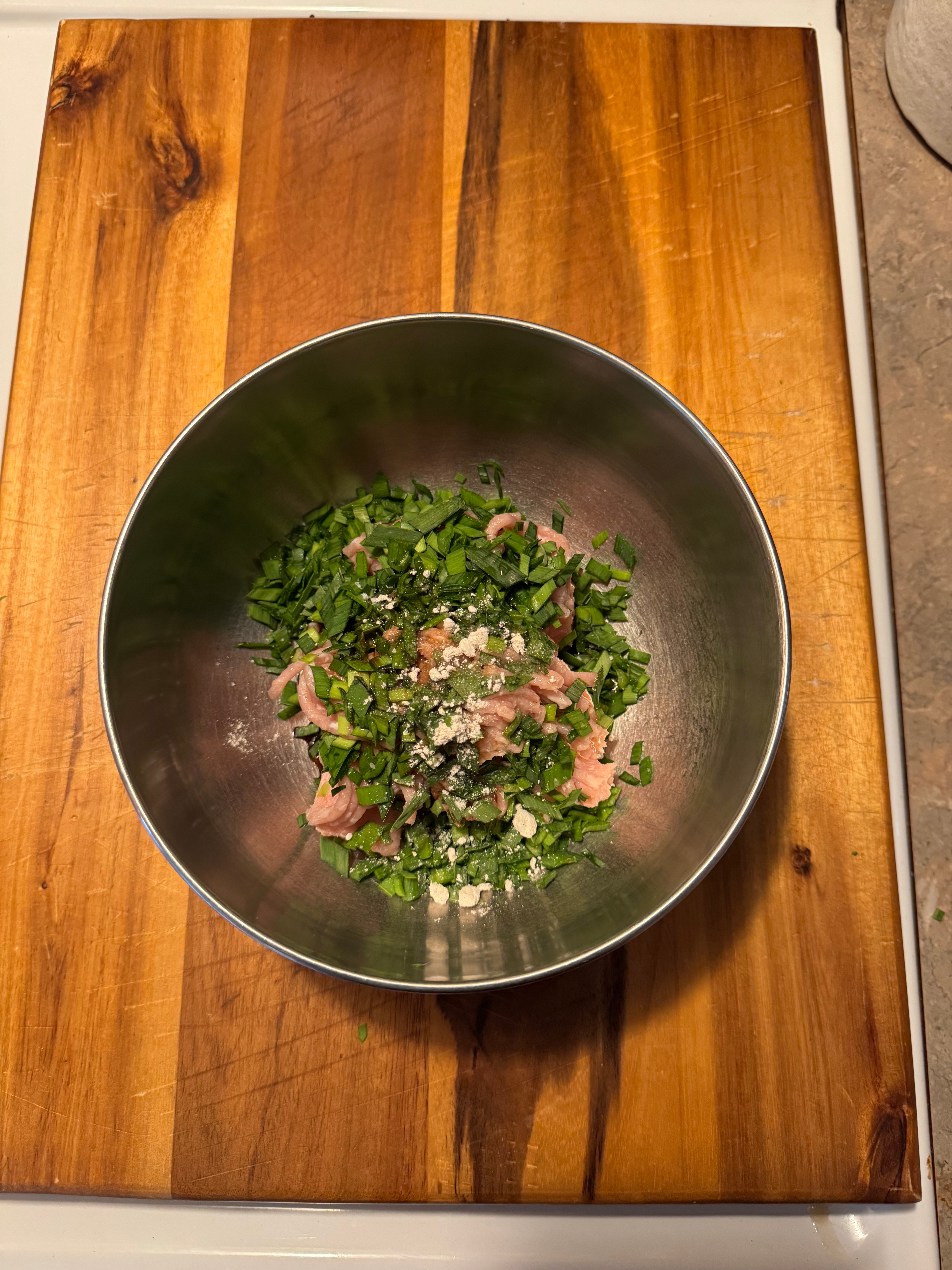 Ingredients in a metal bowl on a wooden surface, including chopped herbs, sliced chicken, and spices