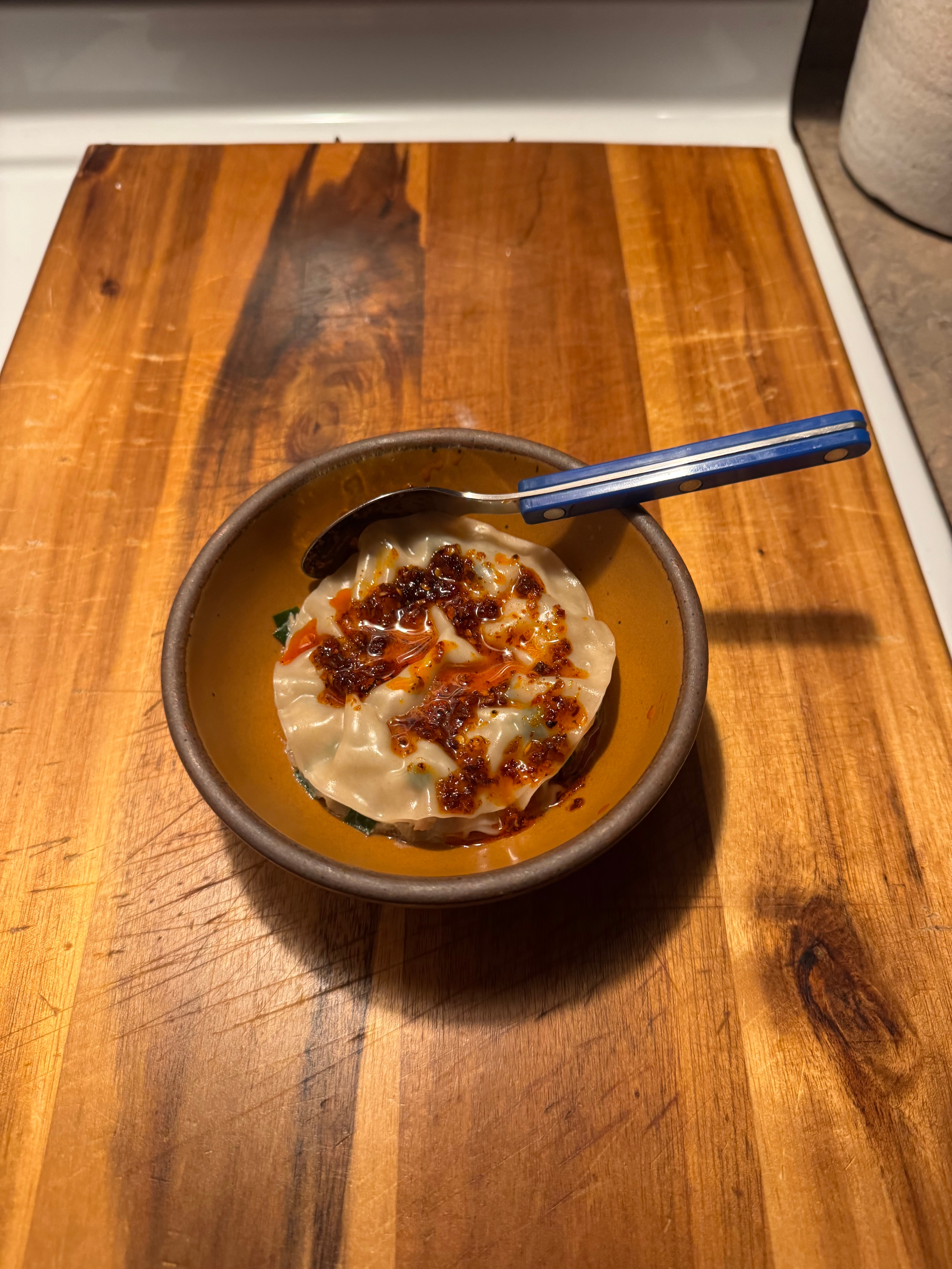 A bowl of dumplings topped with chili oil on a wooden cutting board, with a blue-handled spoon resting on the bowl's edge