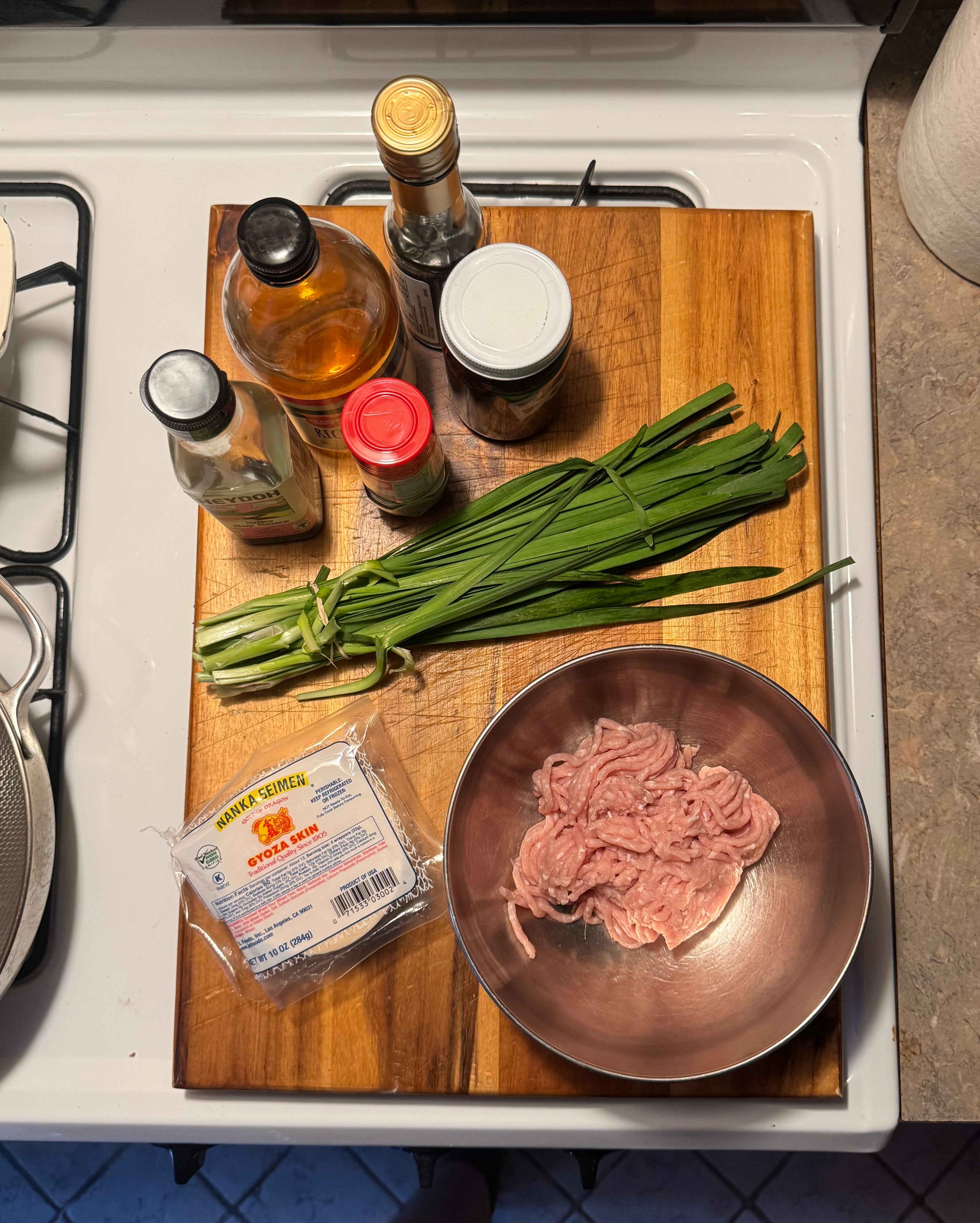 Chopping board with ingredients: green onions, ground meat in a bowl, various sauces, and a tofu package on a kitchen counter