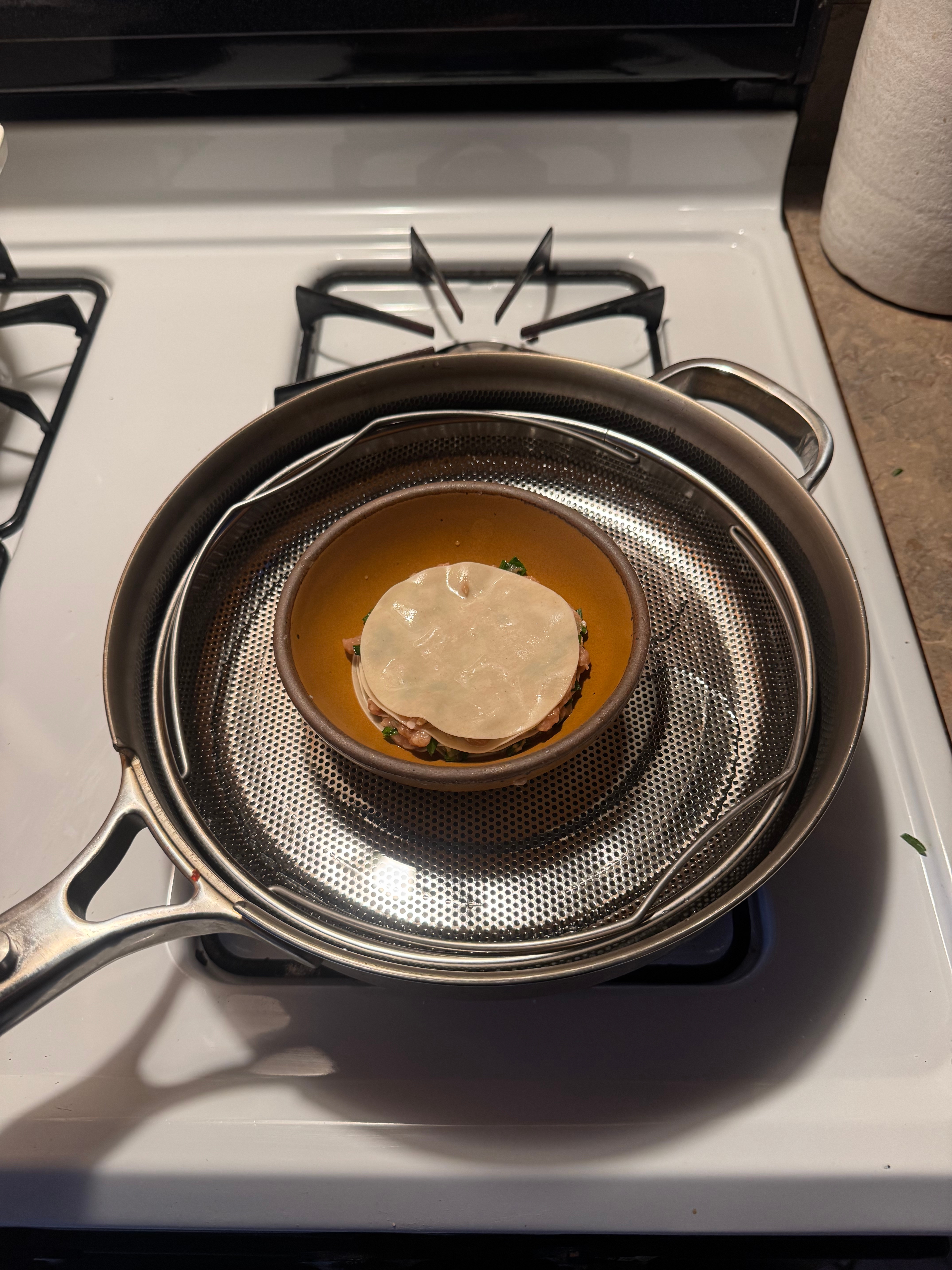 A pan on a stove with a stainless steel steamer basket holding a small dish, likely for cooking or steaming food