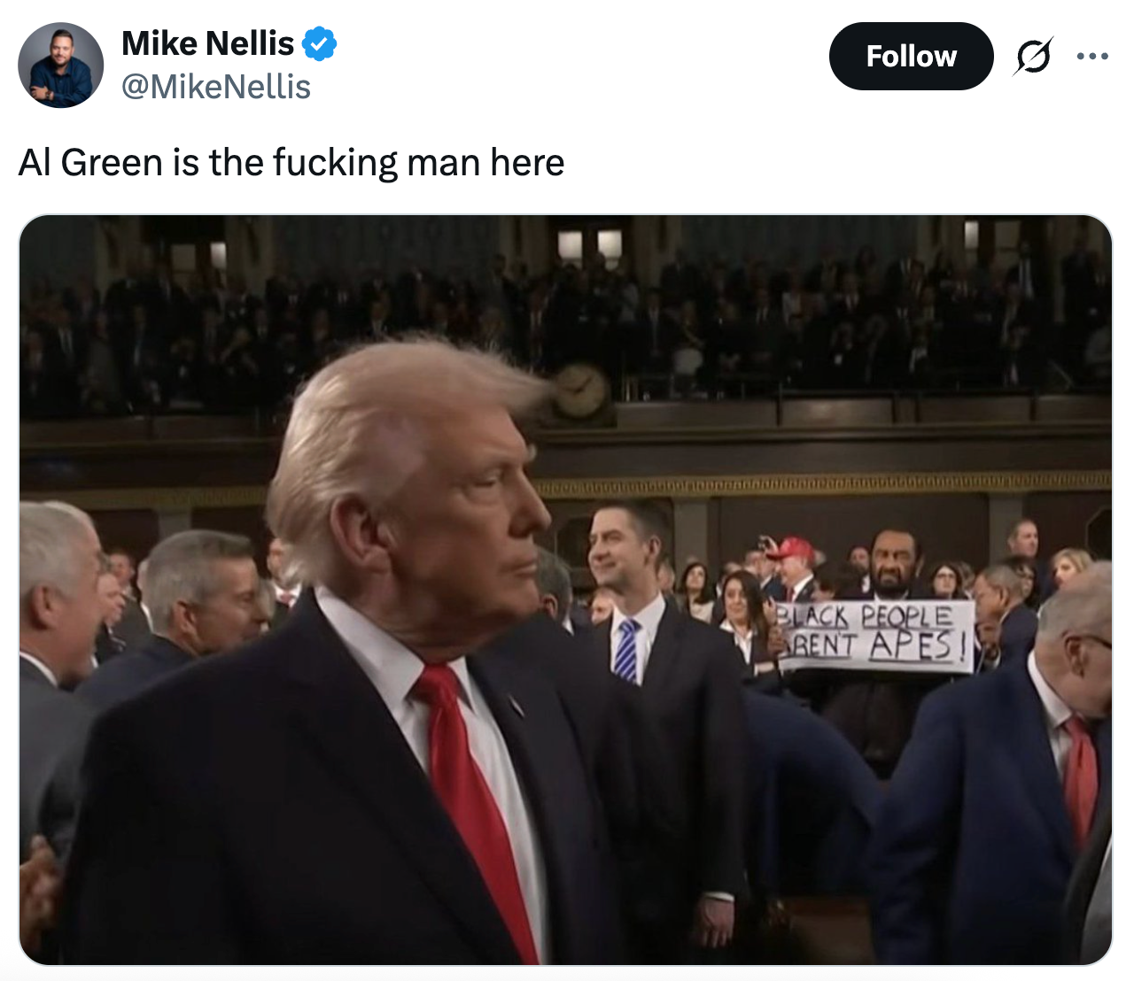 En mann i dress med rødt slips er i forgrunnen. I bakgrunnen holder en person et skilt der det står: 