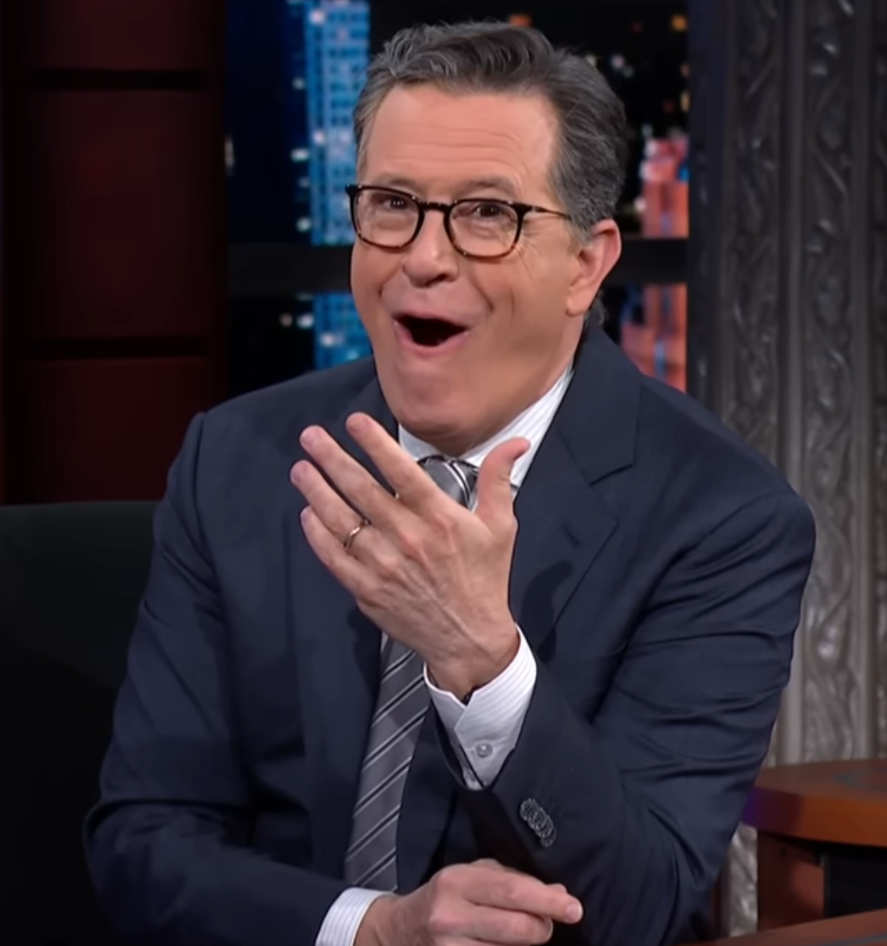 Stephen Colbert in a suit and tie is seated, with an expressive, surprised look, and hand raised near their face on a talk show set