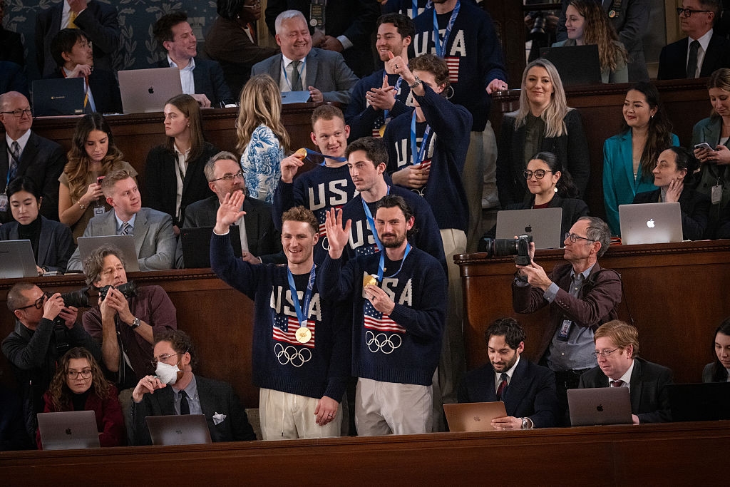 Gruppe mennesker iført matchende OL-gensere i USA, noen vinker. De står i et overfylt rom med andre som sitter og bruker bærbare datamaskiner