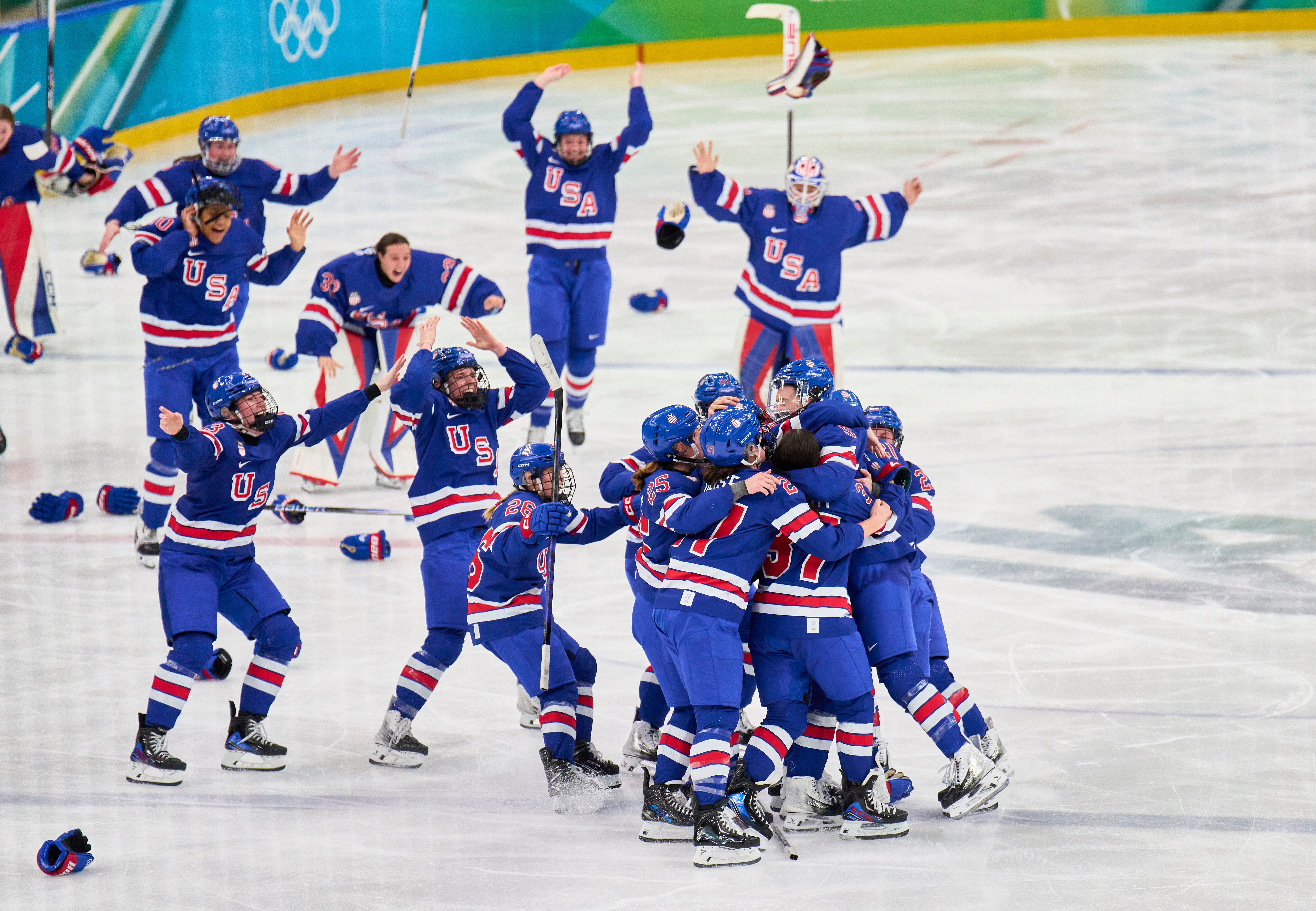 USAs hockeylag for kvinner feirer seier på skøytebane, spillere omfavner og jubler med løftede armer. Hockeysticks og hjelmer spredt rundt