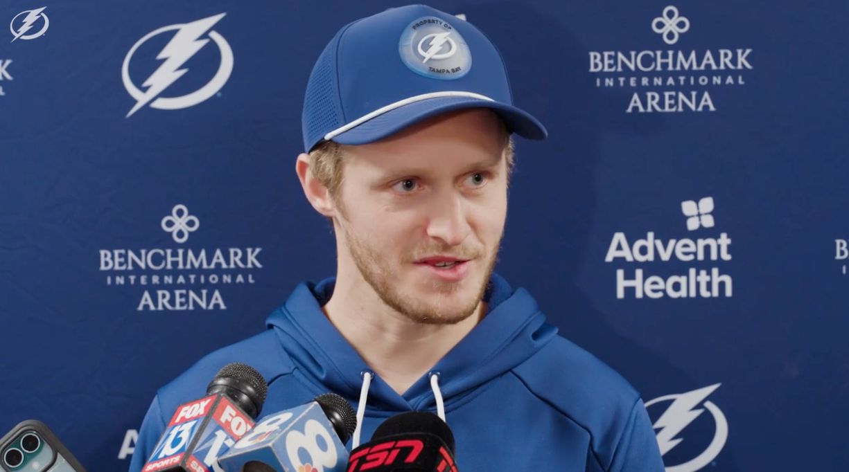 En person i blå hettegenser og caps snakker til pressen på en sportsarena i bakgrunnen. Mikrofoner fra ulike medier er foran