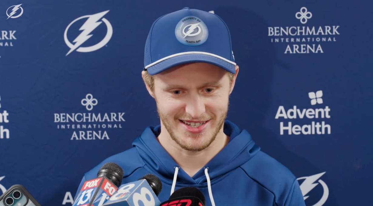 En hockeyspiller iført laghette og hettegenser snakker på en pressebegivenhet med flere mikrofoner foran seg