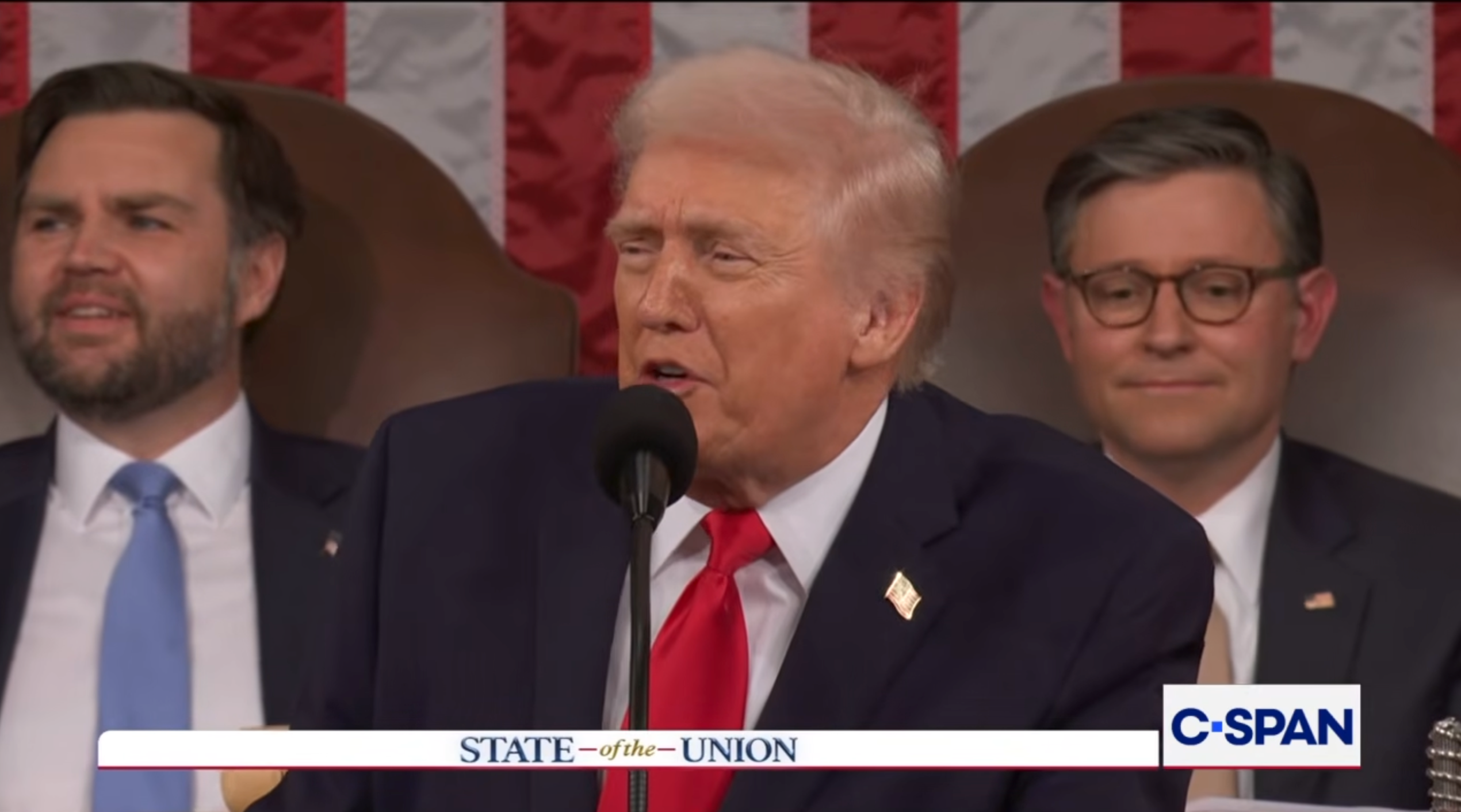 En mann i dress snakker på et podium med «State of the Union»; banner; to menn sitter bak ham