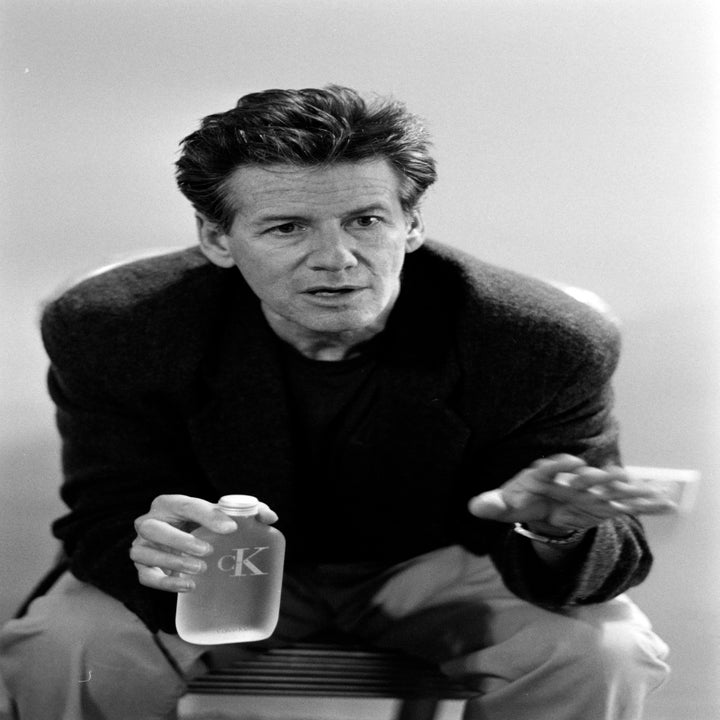 A man sits with a relaxed posture, wearing a casual jacket and holding a bottle with "cK" visible. His expression is thoughtful