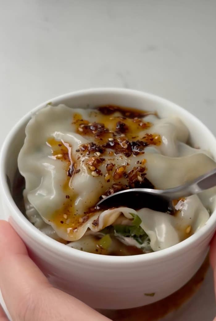 A bowl of dumplings topped with spicy chili oil, with someone holding a spoon ready to take a bite