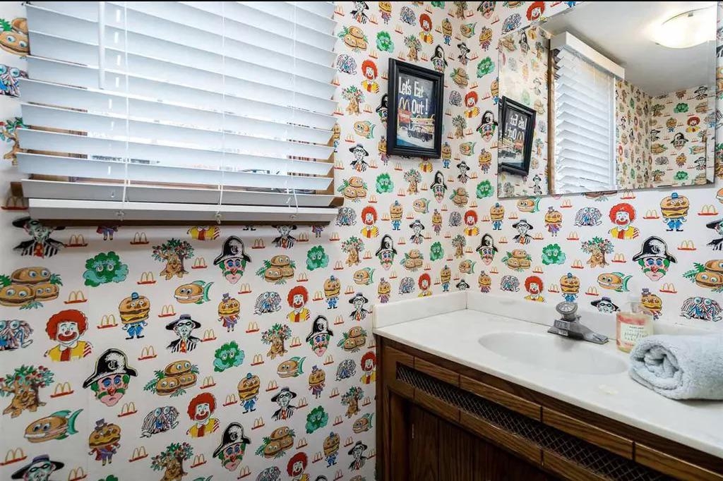 Bathroom with McDonald's-themed wallpaper featuring assorted  characters, a tiny  partition  mirror, woody  cabinet, framed paper   connected  the wall, and a towel connected  the sink