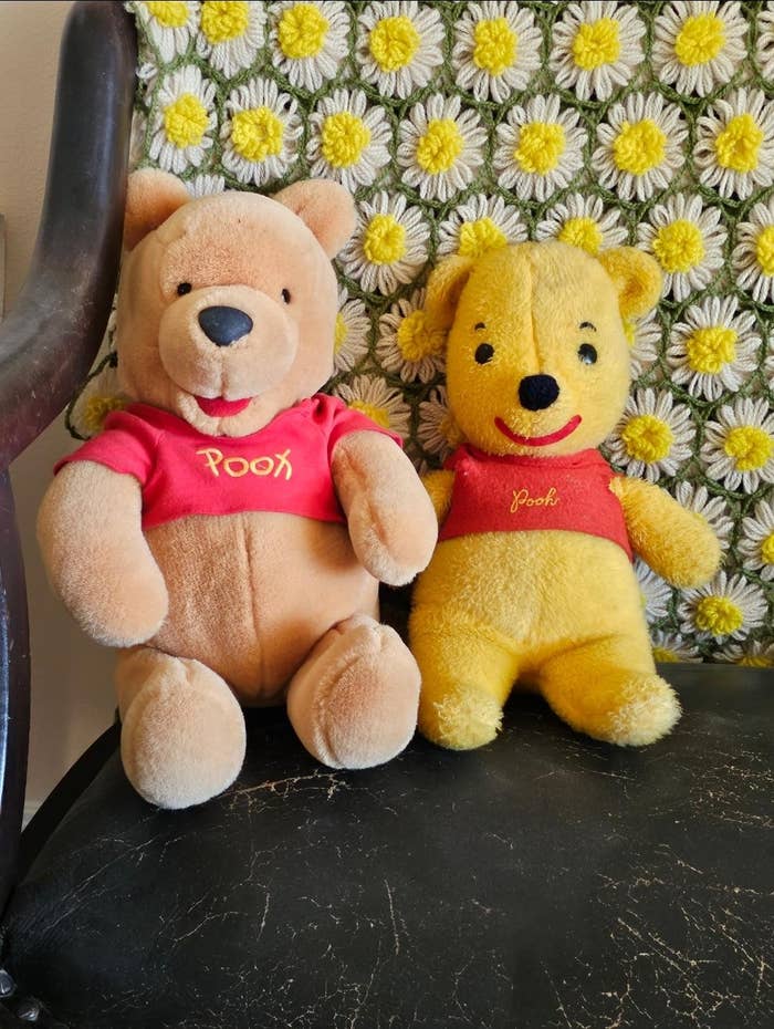 Two plush Pooh carnivore  toys connected  a chair. The near  carnivore  has a much  modern   design, portion    the close    carnivore  looks vintage. They some  deterioration  reddish  shirts with "Pooh" text