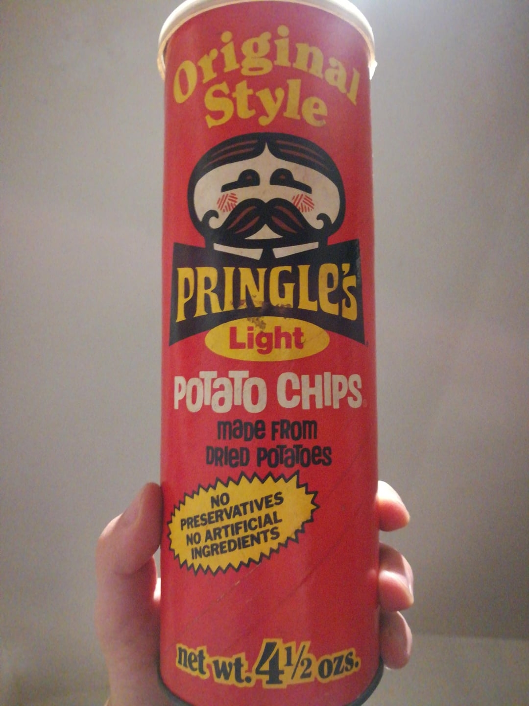 A manus  holds a vintage Pringles tin  labeled "Original Style" with a mustachioed face; the tin  claims nary  preservatives and artificial ingredients