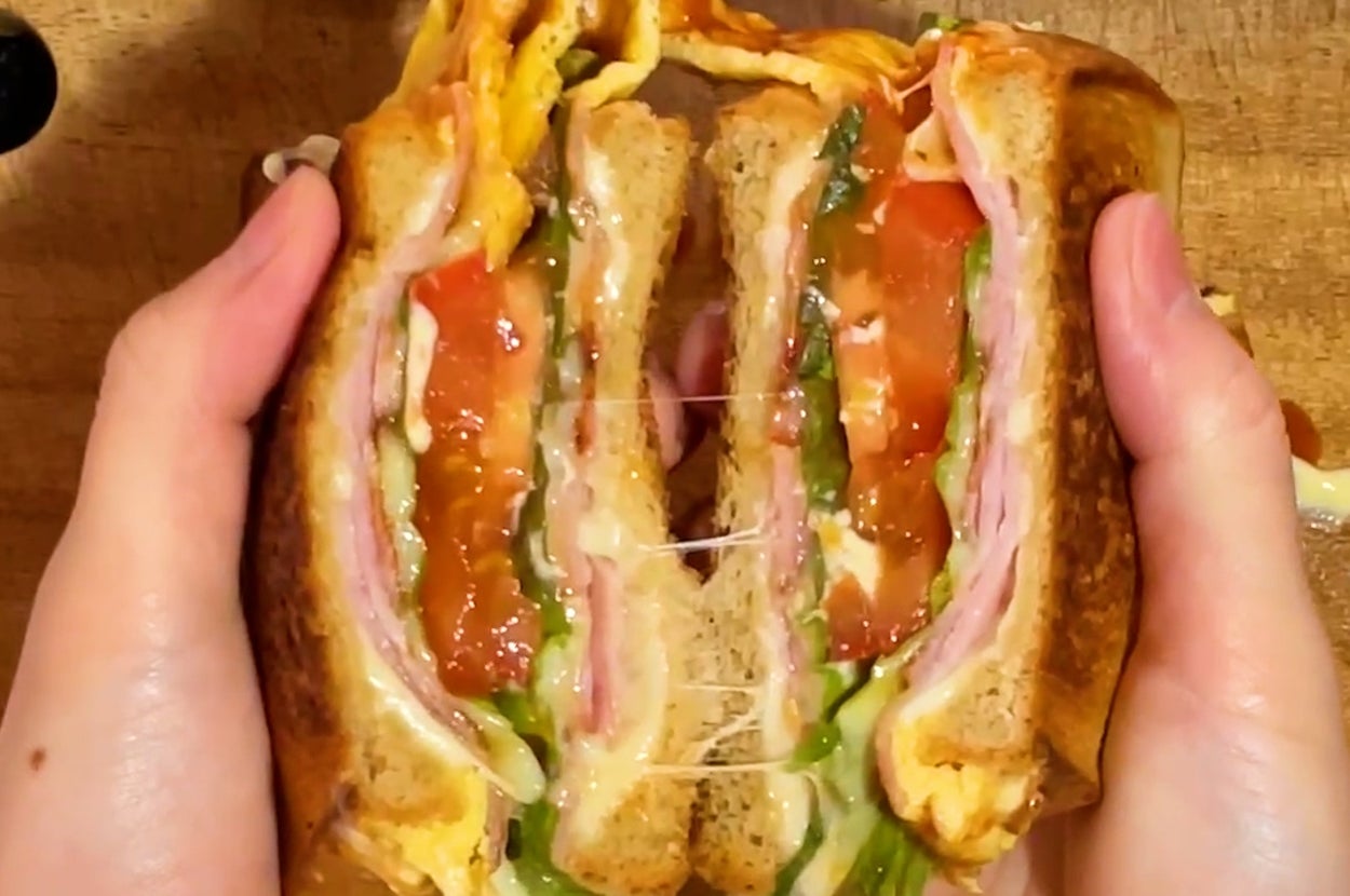 サンドイッチを持つ手。トマト、レタス、ハム、溶けたチーズが層になっている。