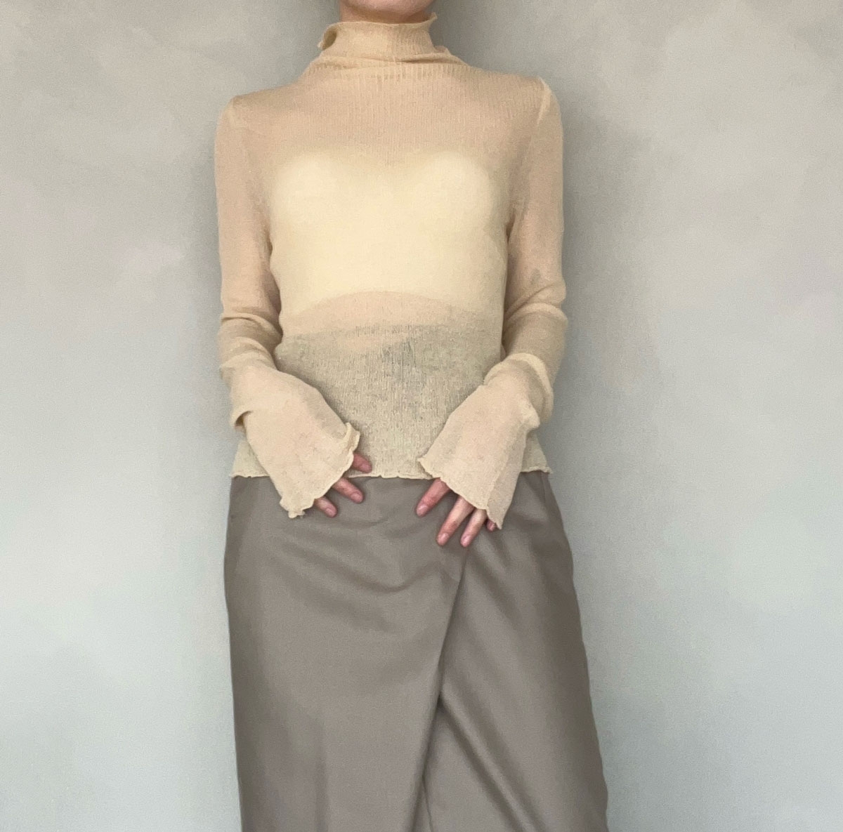 Person wearing a sheer turtleneck top with unique sleeve cuffs and a long, fitted skirt standing against a plain backdrop
