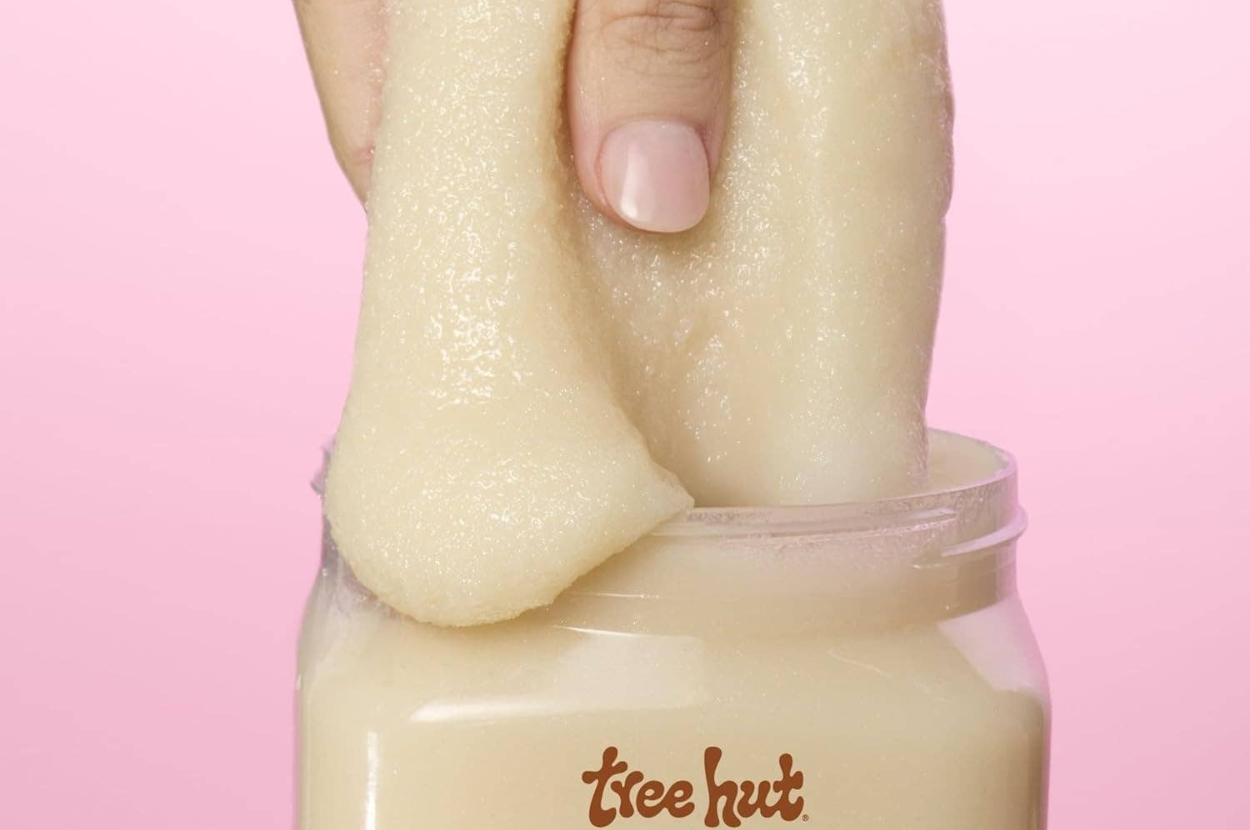 Hand dipping into a jar of Tree Hut body scrub against a pink background, highlighting texture for exfoliation in a shopping context